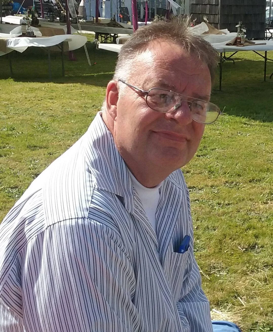 A man wearing glasses and a striped shirt, sitting outdoors on grass, smiling at the camera with tables and chairs in the background.