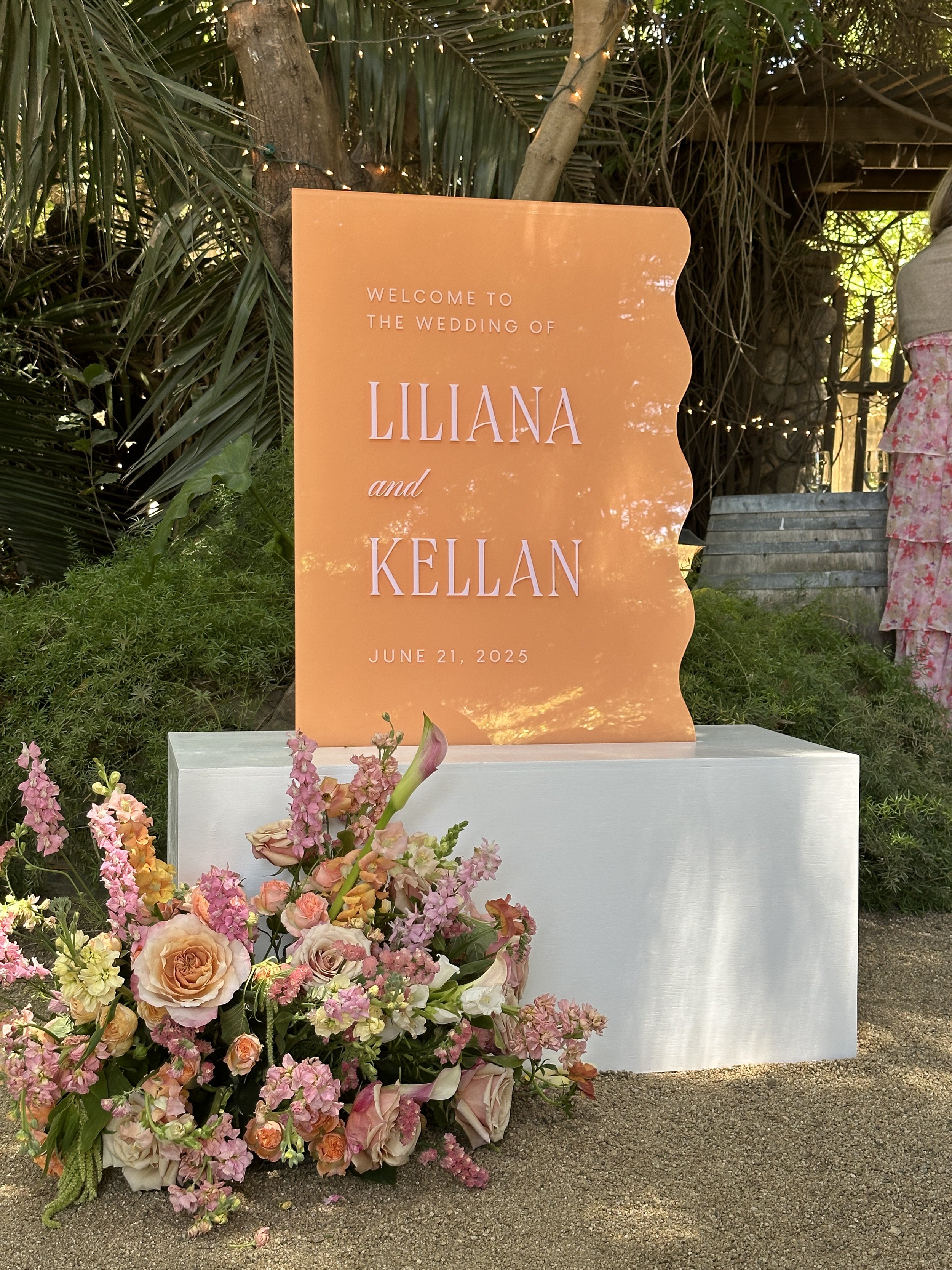 A whimsical peach and pink wavy welcome sign for a wedding at Holly Farm in Carmel, California. The welcome sign is displayed on a white box base with lush florals placed in front of the display.