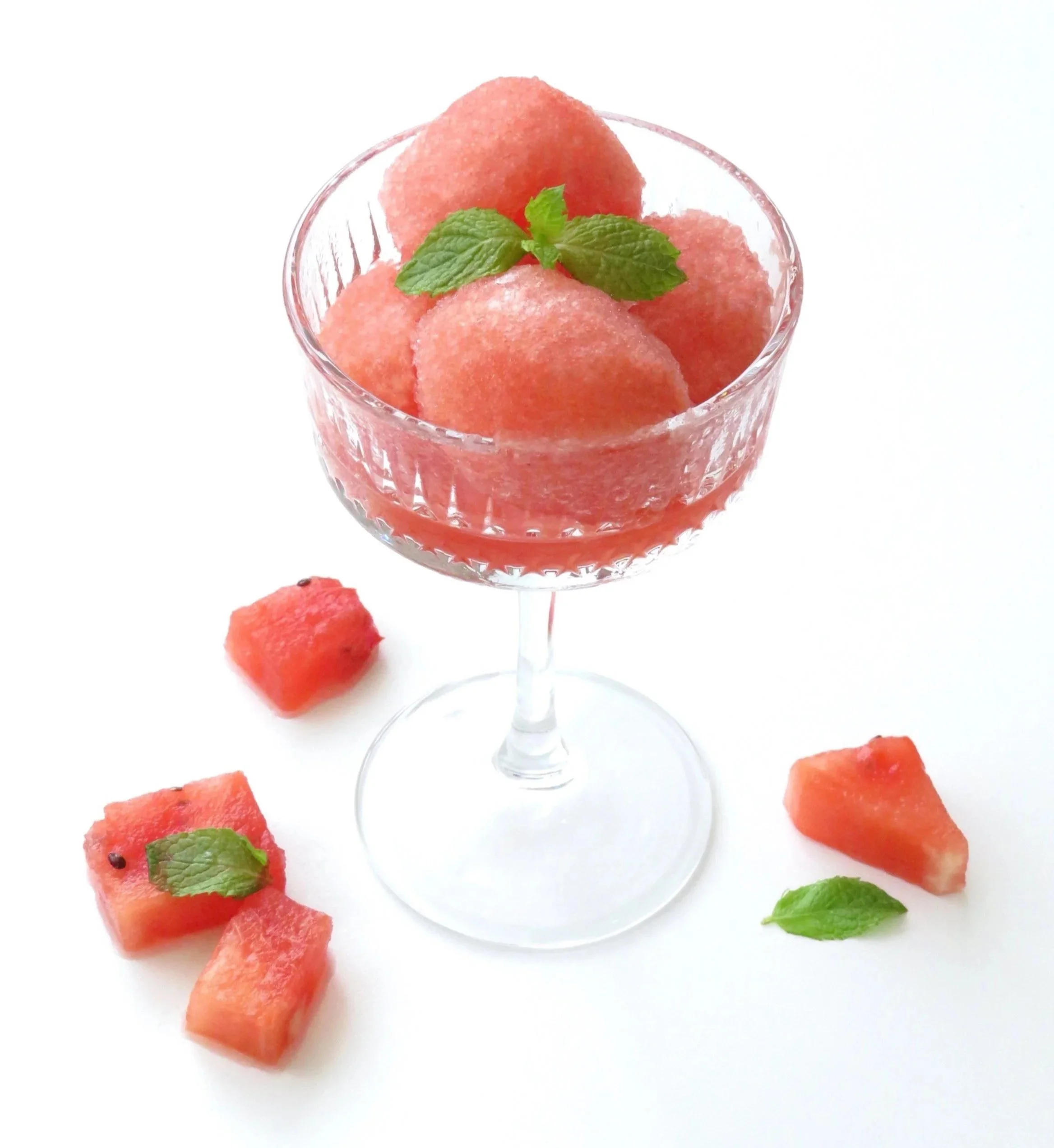 Scoops of watermelon sorbet in serving glass bowl with mint garnish and watermelon cubes scattered around