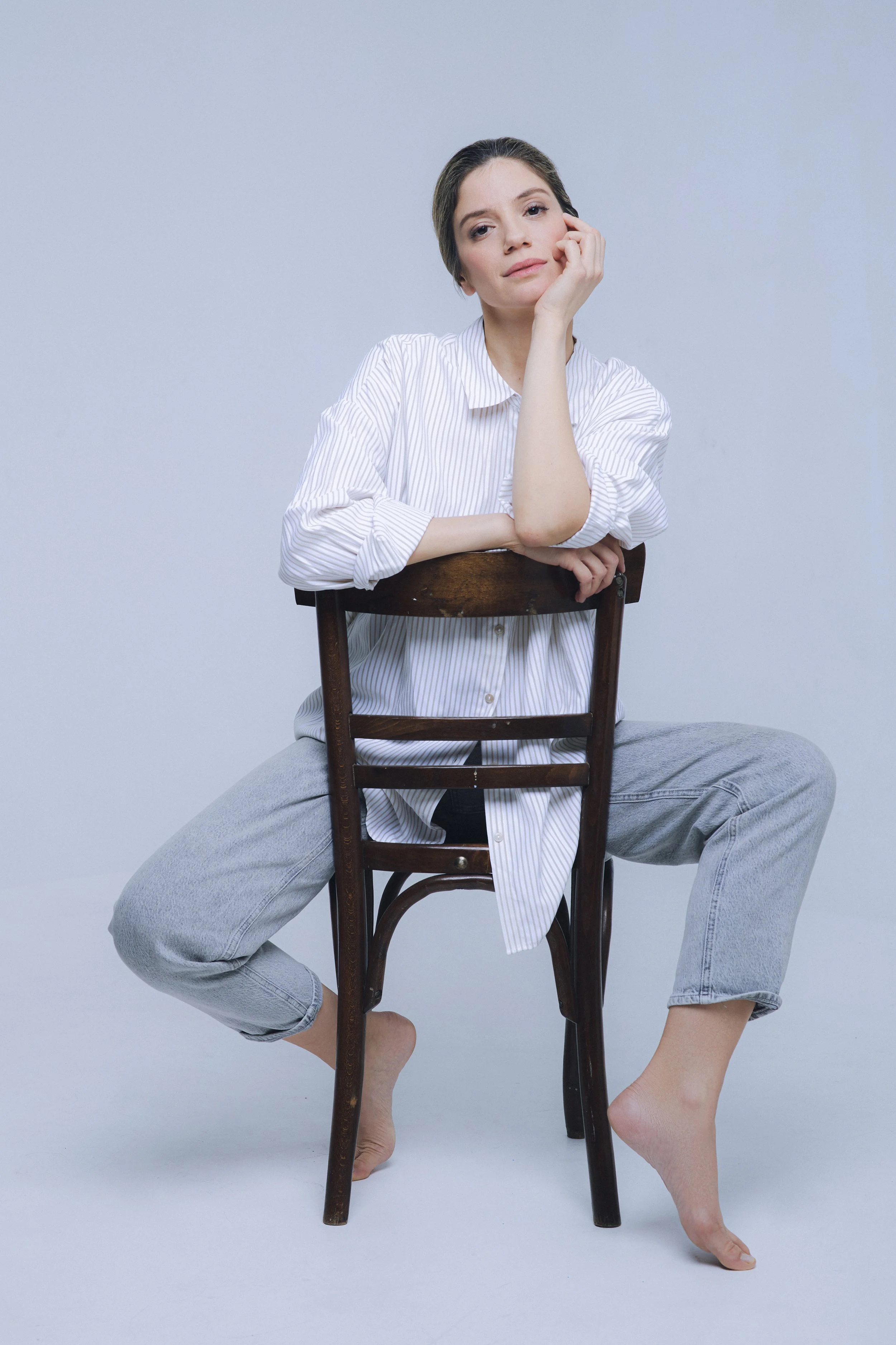 A woman sitting on a wooden chair, barefoot, wearing a white striped shirt and gray jeans, with her left arm resting on the back of the chair and her chin resting on her left hand, looking at the camera with a neutral expression.