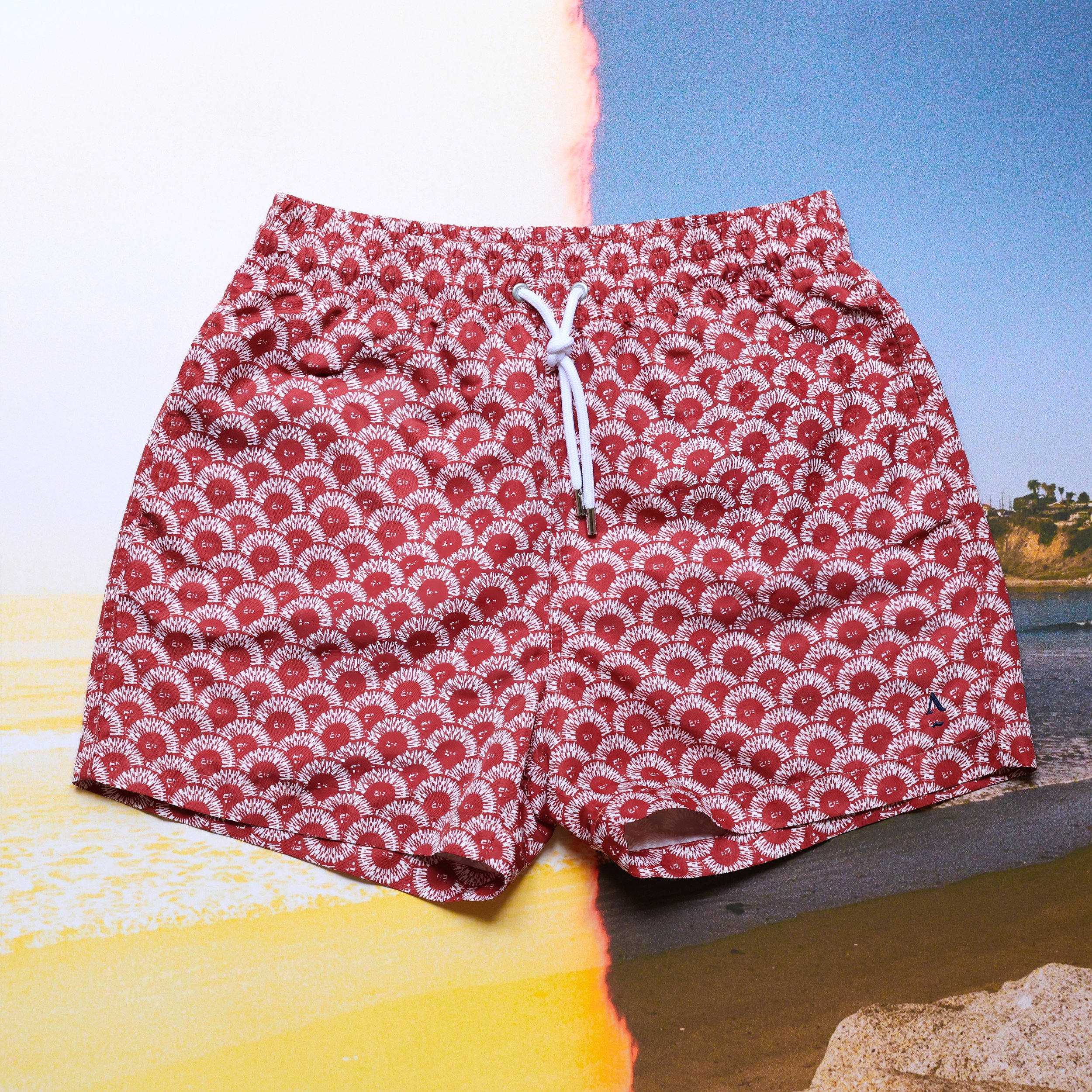Red and white floral patterned swim trunks with a white drawstring, placed on a beach background.