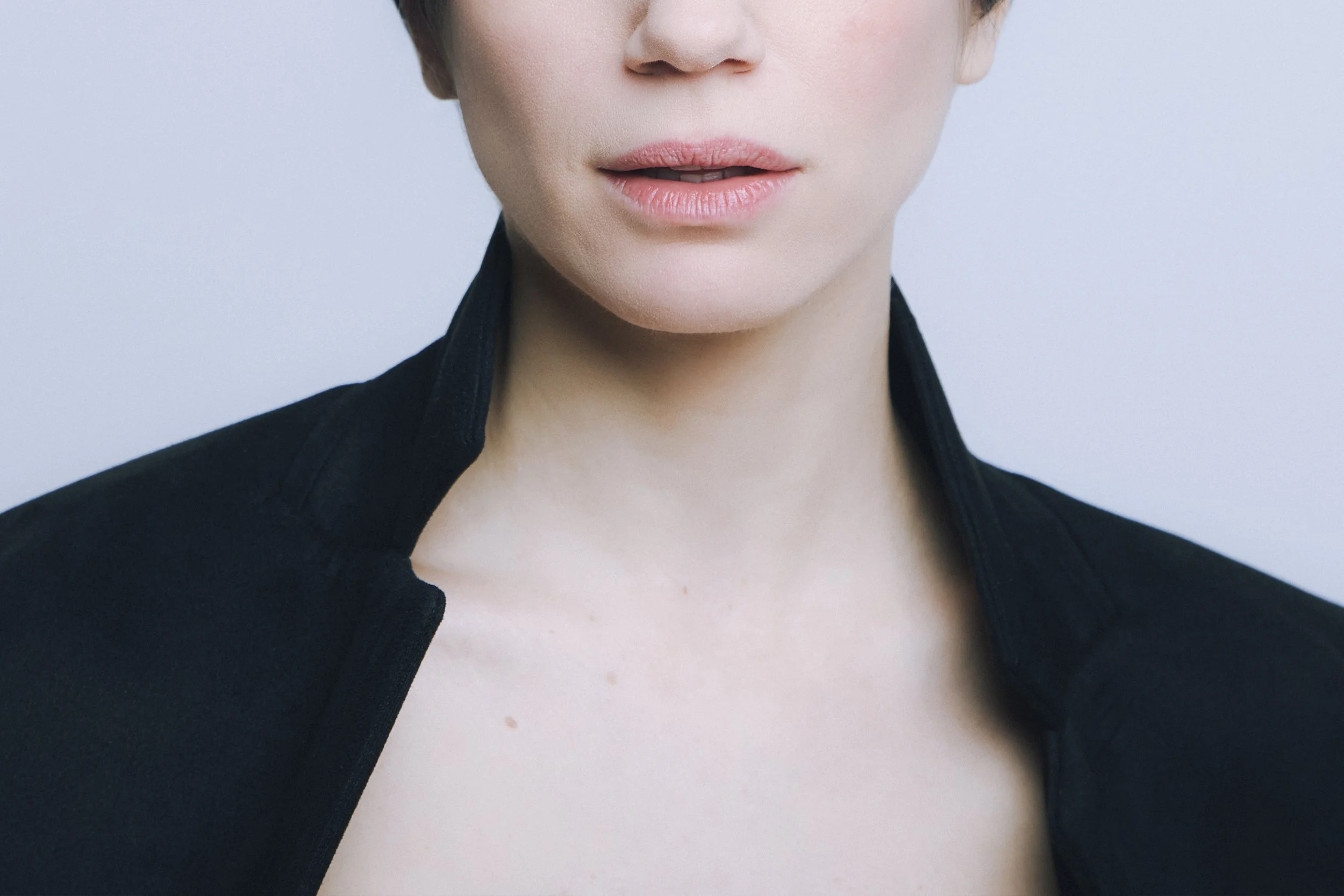 Close-up of a woman with short hair and fair skin, wearing a black jacket with the collar slightly turned up, against a plain light background.