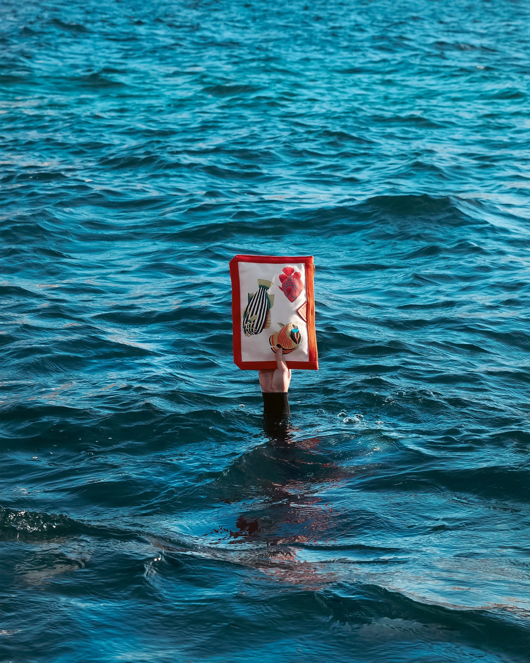 A hand holding a colorful fabric item above the water, with the hand emerging from the water, in a body of water that appears to be the ocean.
