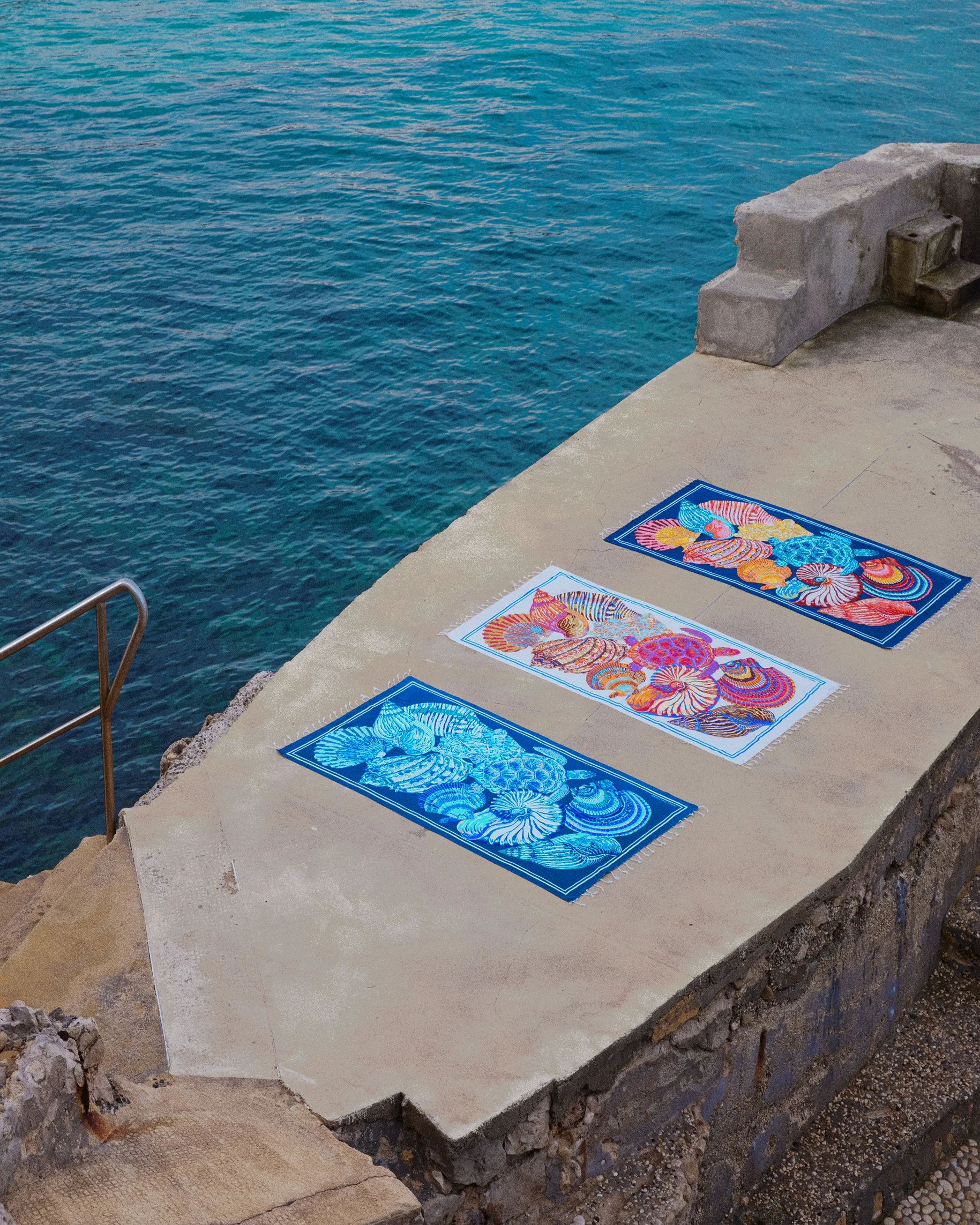 Three colorful beach towels with sea shell patterns are laid out on a concrete pier beside the water, with steps leading into the water in the background.