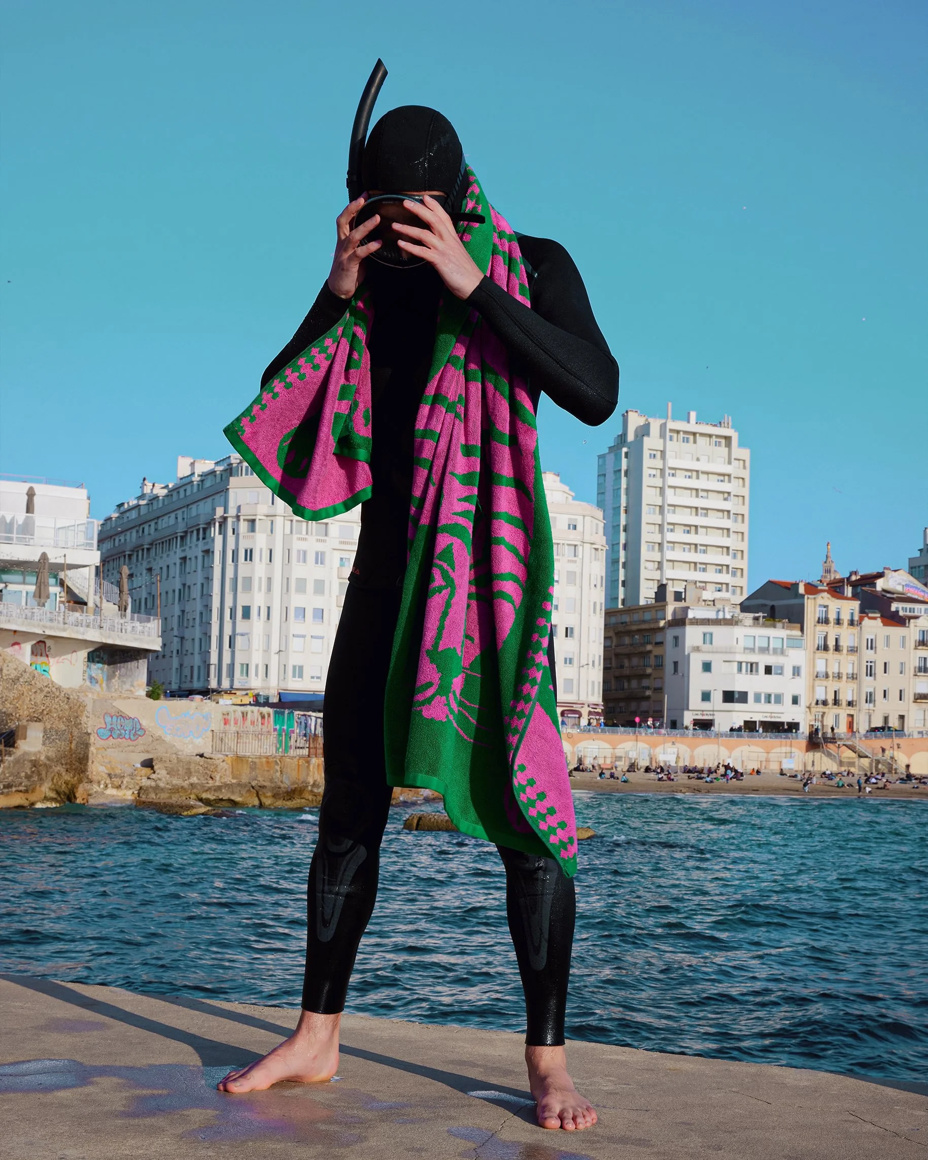 A person in a wetsuit, holding a snorkeling mask and snorkel, standing barefoot on a pier near the water, with buildings and a beach with people in the background.
