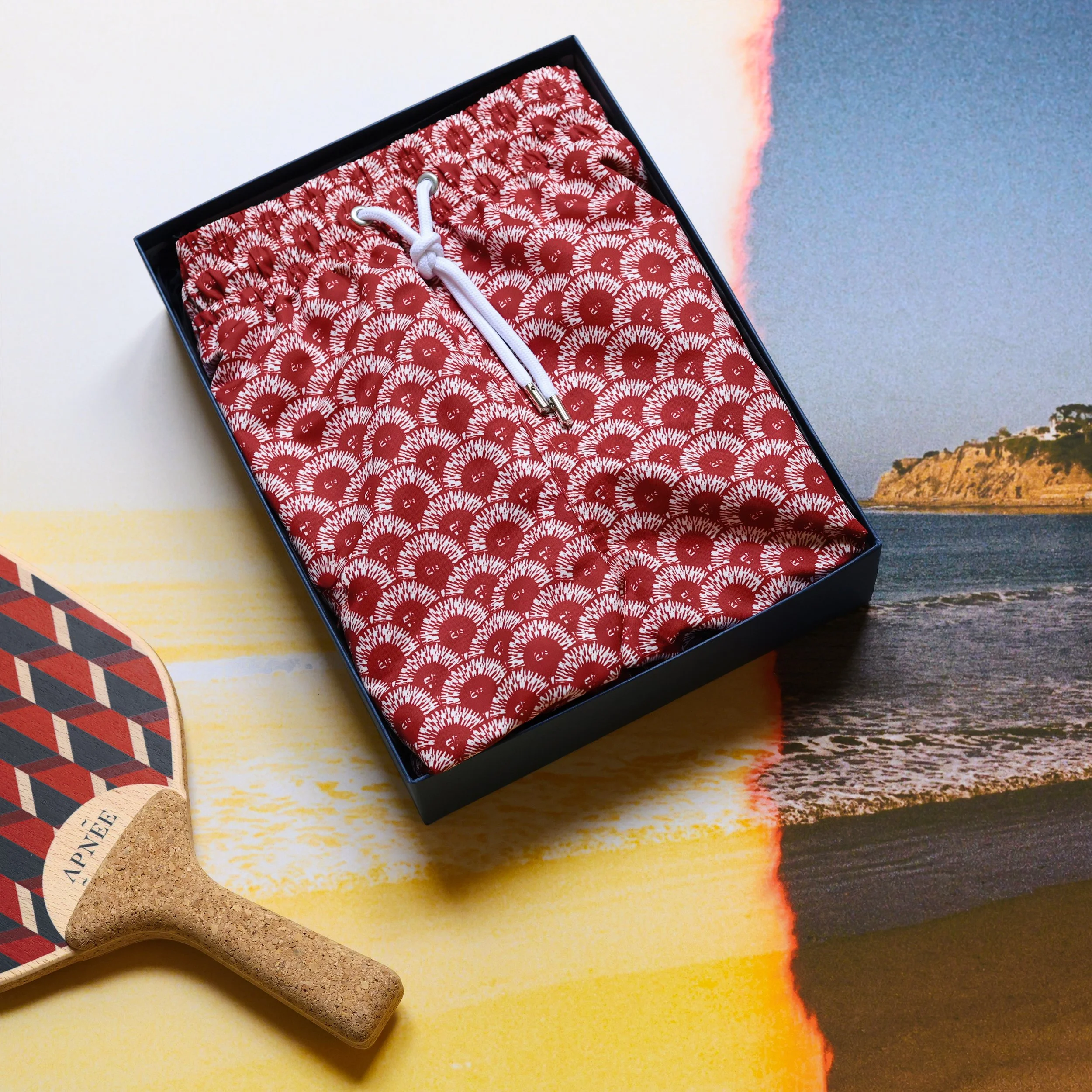 A gift box containing red and white swim trunks with a drawstring, placed on a colorful board game with a paddle-shaped game piece nearby, on a table with a beach scene background.
