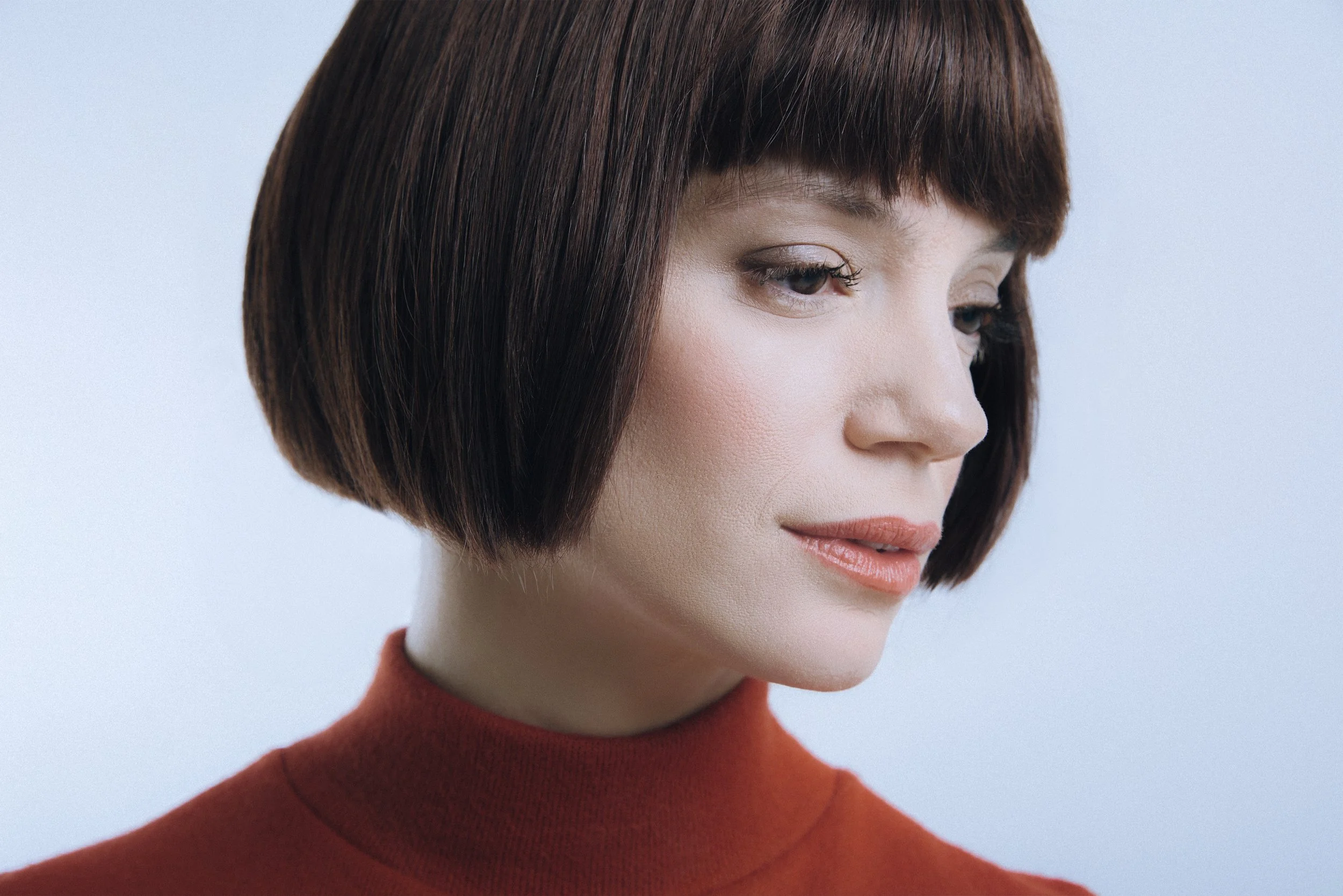Close-up of a woman with short brown hair wearing a red turtleneck, looking down thoughtfully.