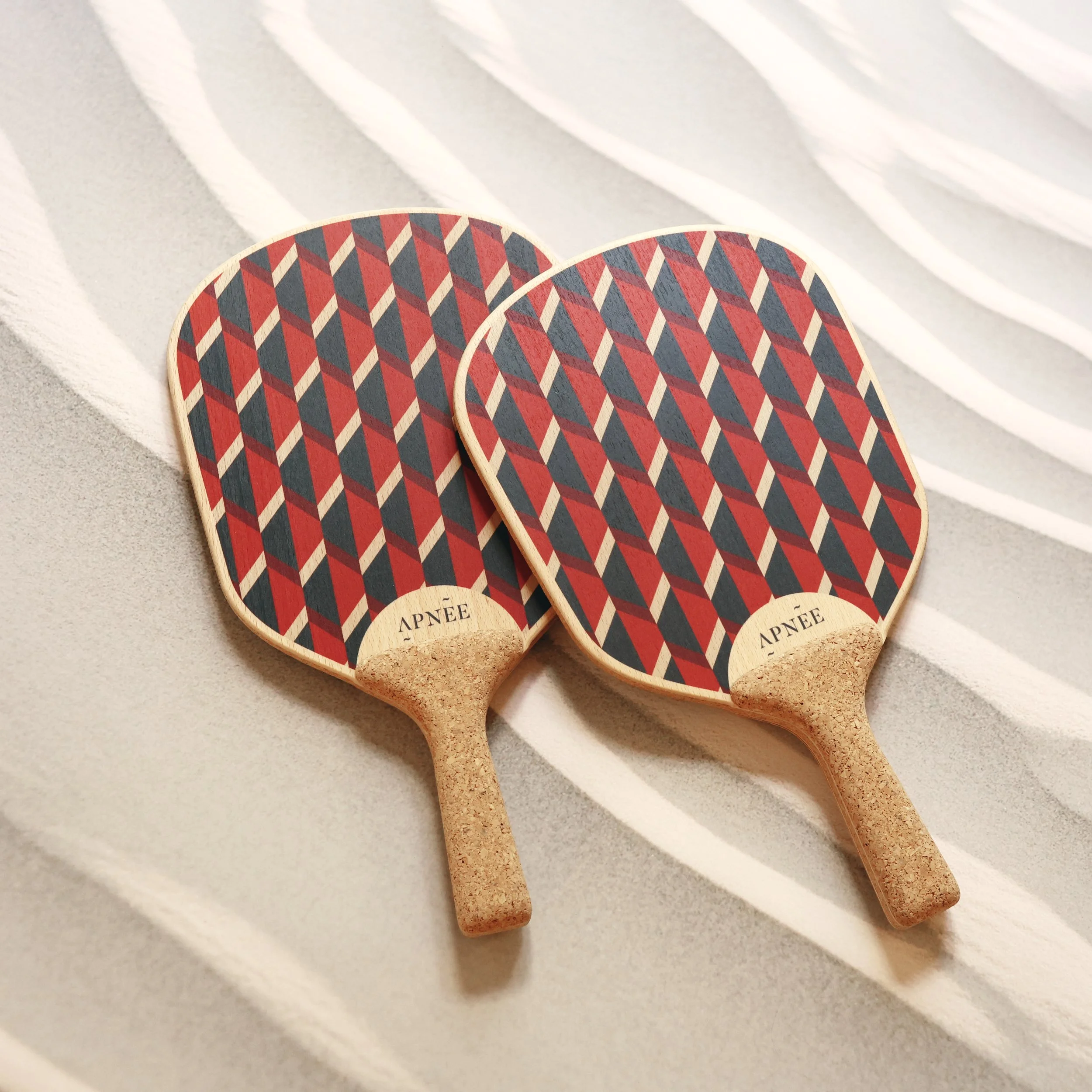 Two ping pong paddles with geometric red, black, and beige pattern on their faces, resting on a textured surface with white ridges.