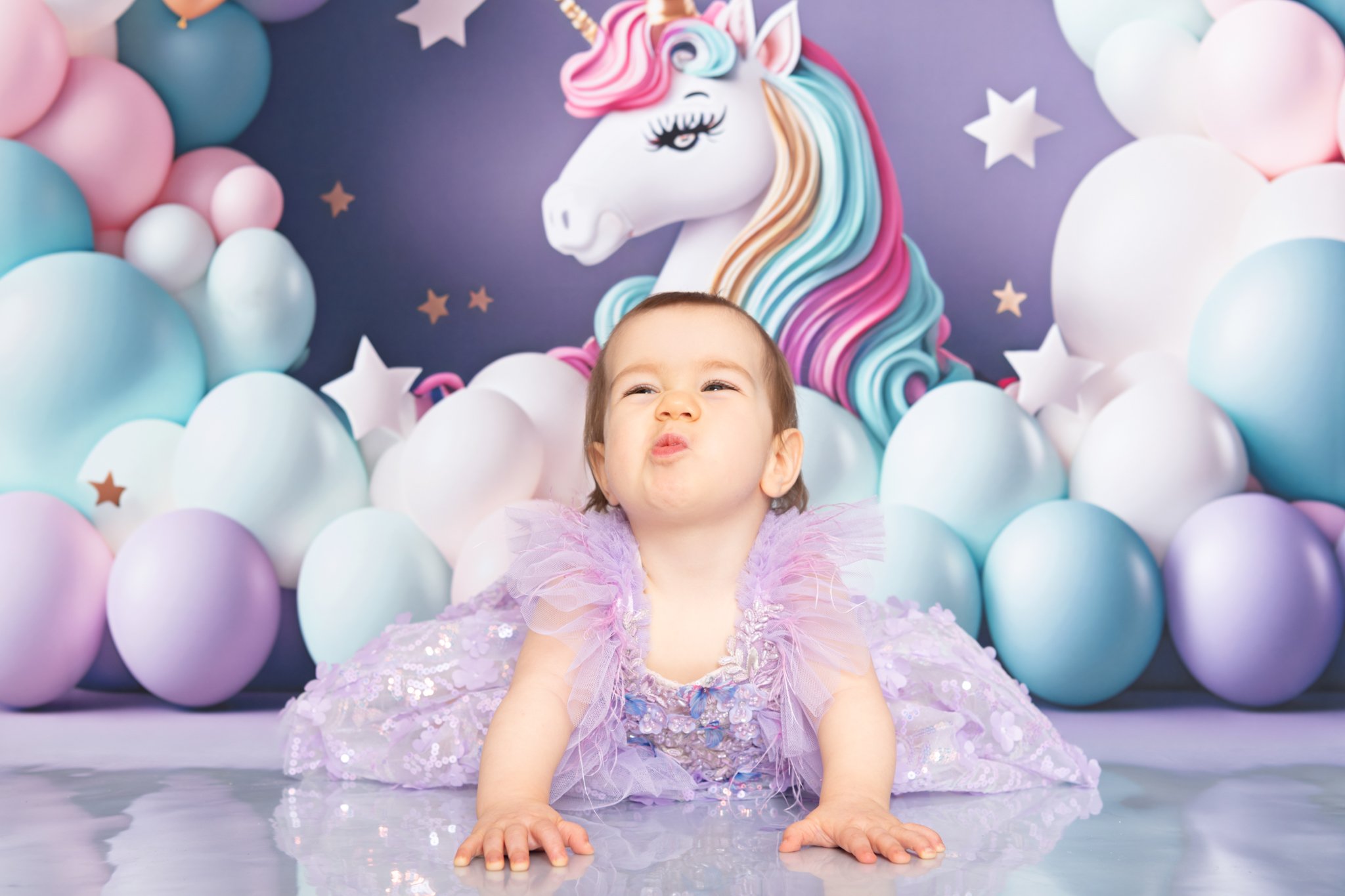 Un bébé en robe violette avec des tulle est assis sur le sol, dans une décoration de fête d'anniversaire avec un thème arc-en-ciel et licorne, comprenant des ballons de différentes couleurs et une licorne en fond.