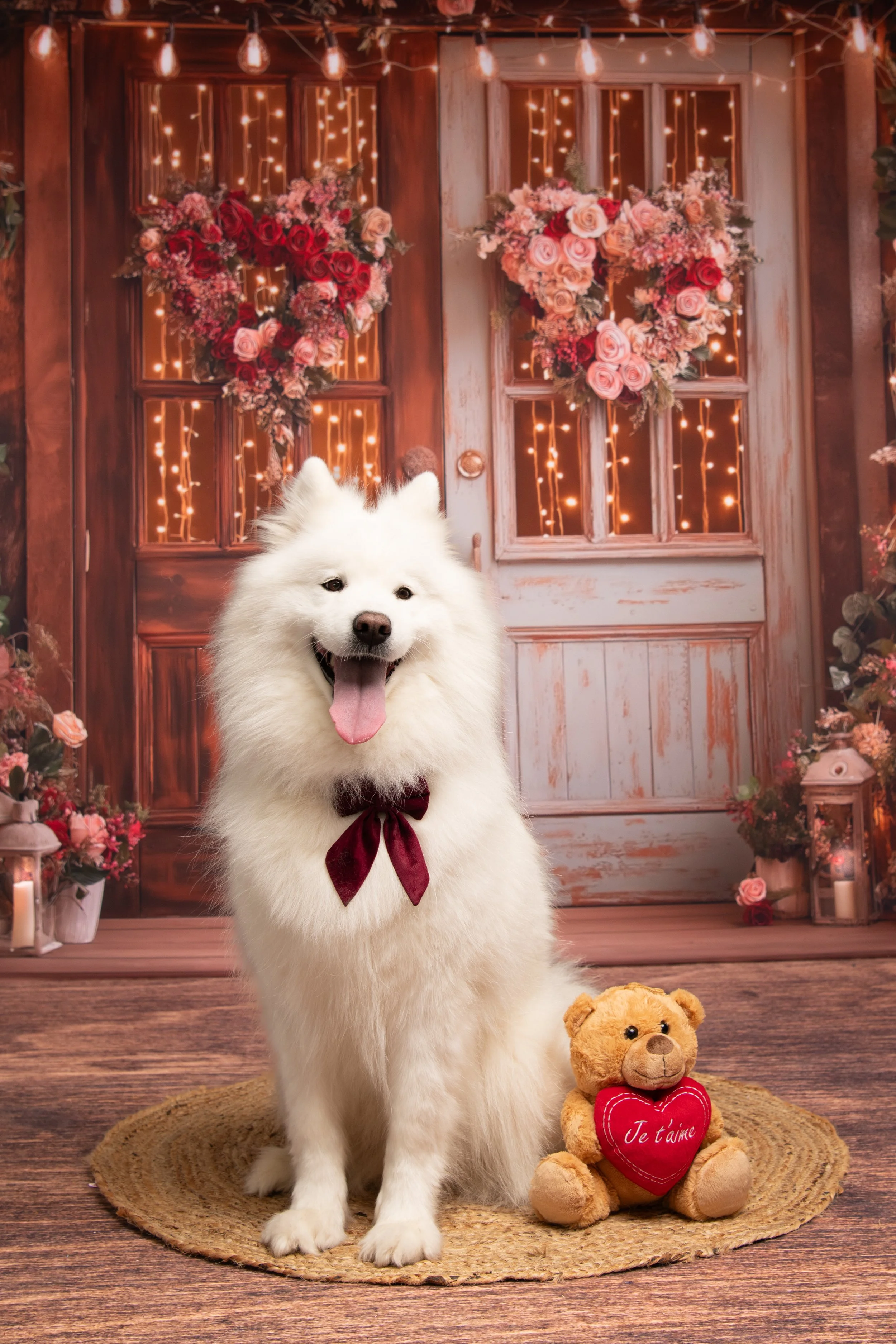 Un chien blanc avec un collier en ruban bordeaux, assis sur un tapis rond en osier, avec un ours en peluche tenant un cœur rouge avec 'Je t'aime' écrit dessus, devant une porte en bois décorée de guirlandes lumineuses et de cœurs en fleurs, ambiance romantique et festive.