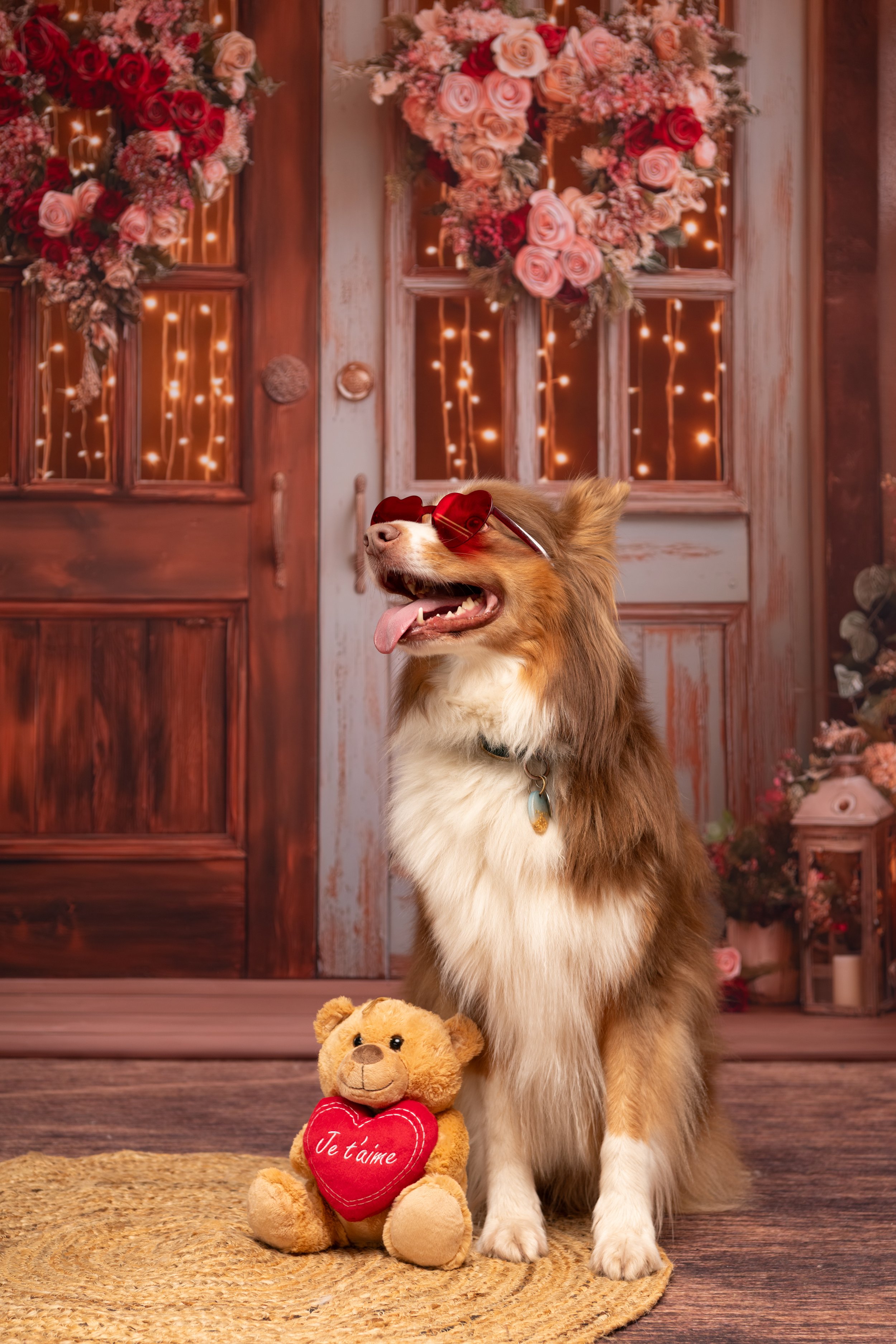 Chien avec des lunettes de soleil rouge assis à côté d'un ours en peluche, devant une porte en bois décorée avec des guirlandes lumineuses et des guirlandes de fleurs, ambiance romantique et chaleureuse.
