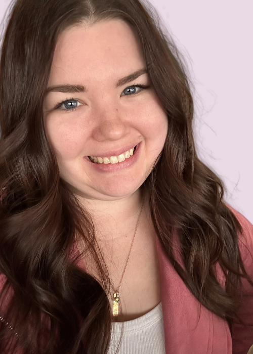 A young woman named Rachel Gunn with long, wavy brown hair, blue eyes, and light skin, smiling and wearing a pink jacket over a white top, against a light background.