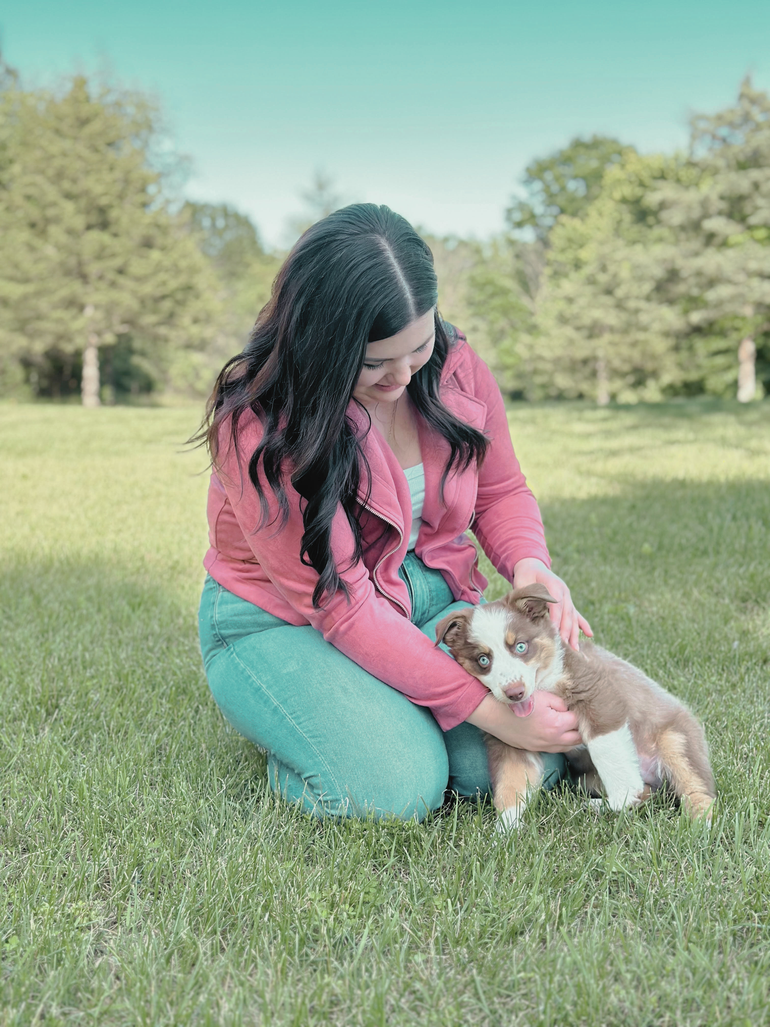 A woman, Rachel Gunn, with long dark hair, wearing a pink jacket and blue jeans, is sitting on grass in a park, playing with a puppy with blue eyes and a white and brown coat.