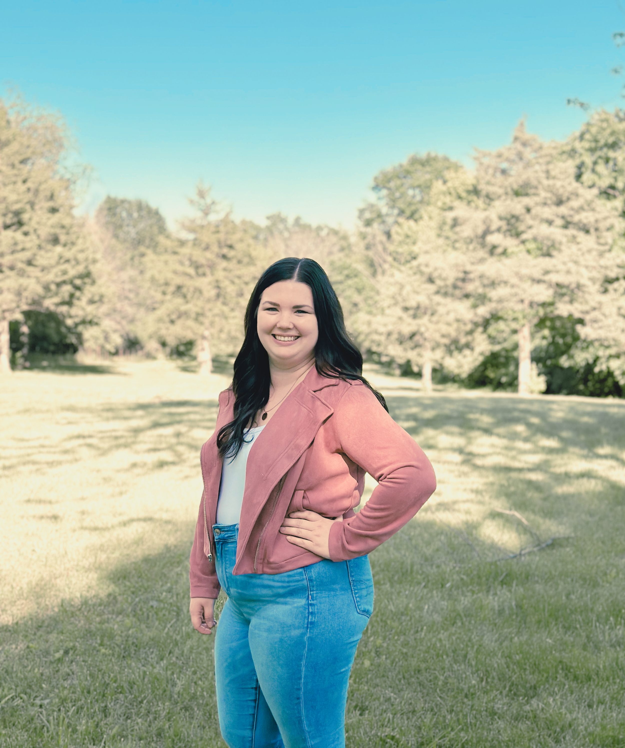 A woman, Rachel Gunn, with long dark hair smiling outdoors in a park, wearing a pink jacket and blue jeans, with trees and grass in the background.