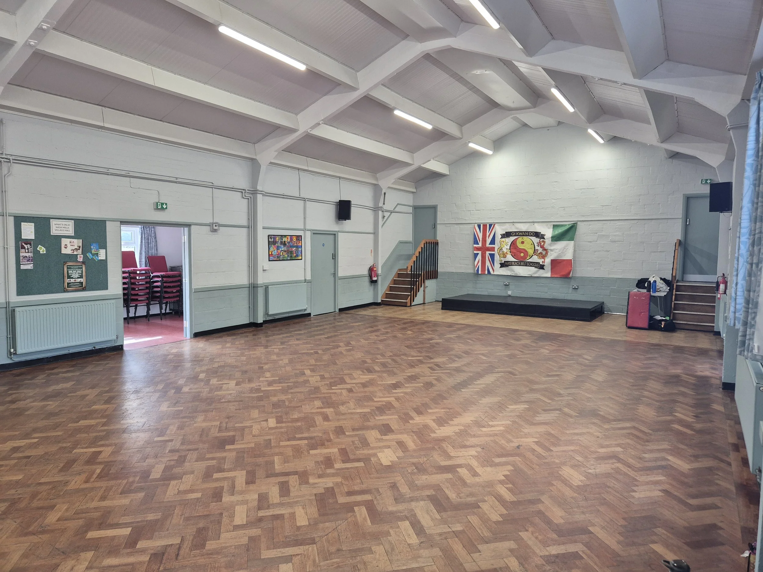 An empty indoor community hall or gymnasium with a wooden floor, light-colored brick walls, ceiling lights, a staircase, banners including one with the British flag and Chinese characters, and various chairs and items stored along the walls.