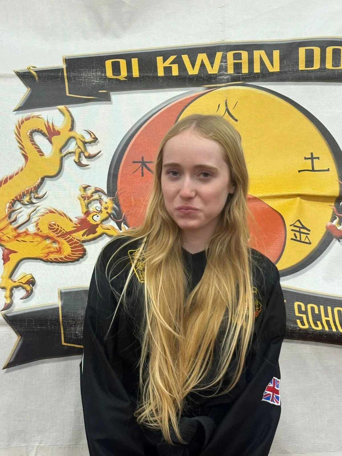 A young woman with long blonde hair wearing a black martial arts uniform with an Union Jack patch on her sleeve. She is standing in front of a Hong Kong-themed background featuring a yellow and red Chinese dragon and a circular emblem with Chinese characters and the words "QI KWAN D" at the top.