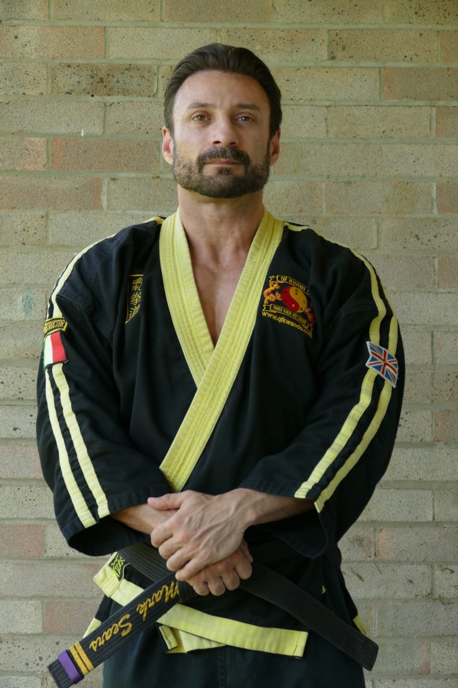 A man with a beard and mustache wearing a black martial arts gi with yellow trim and patches, standing in front of a brick wall, crossing his arms and looking at the camera.