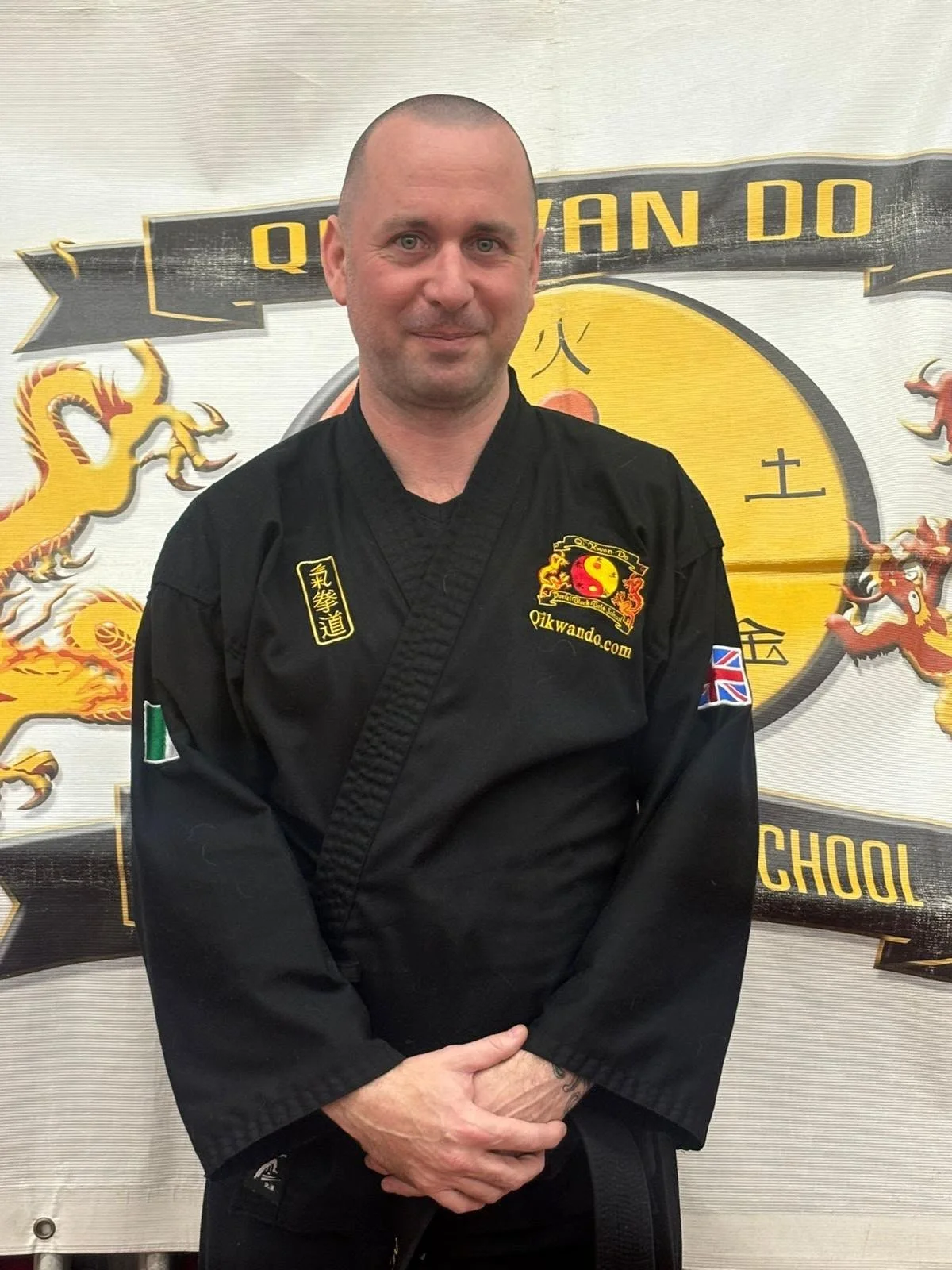A man in a black martial arts uniform standing in front of a yellow and black banner with dragons and Chinese characters.