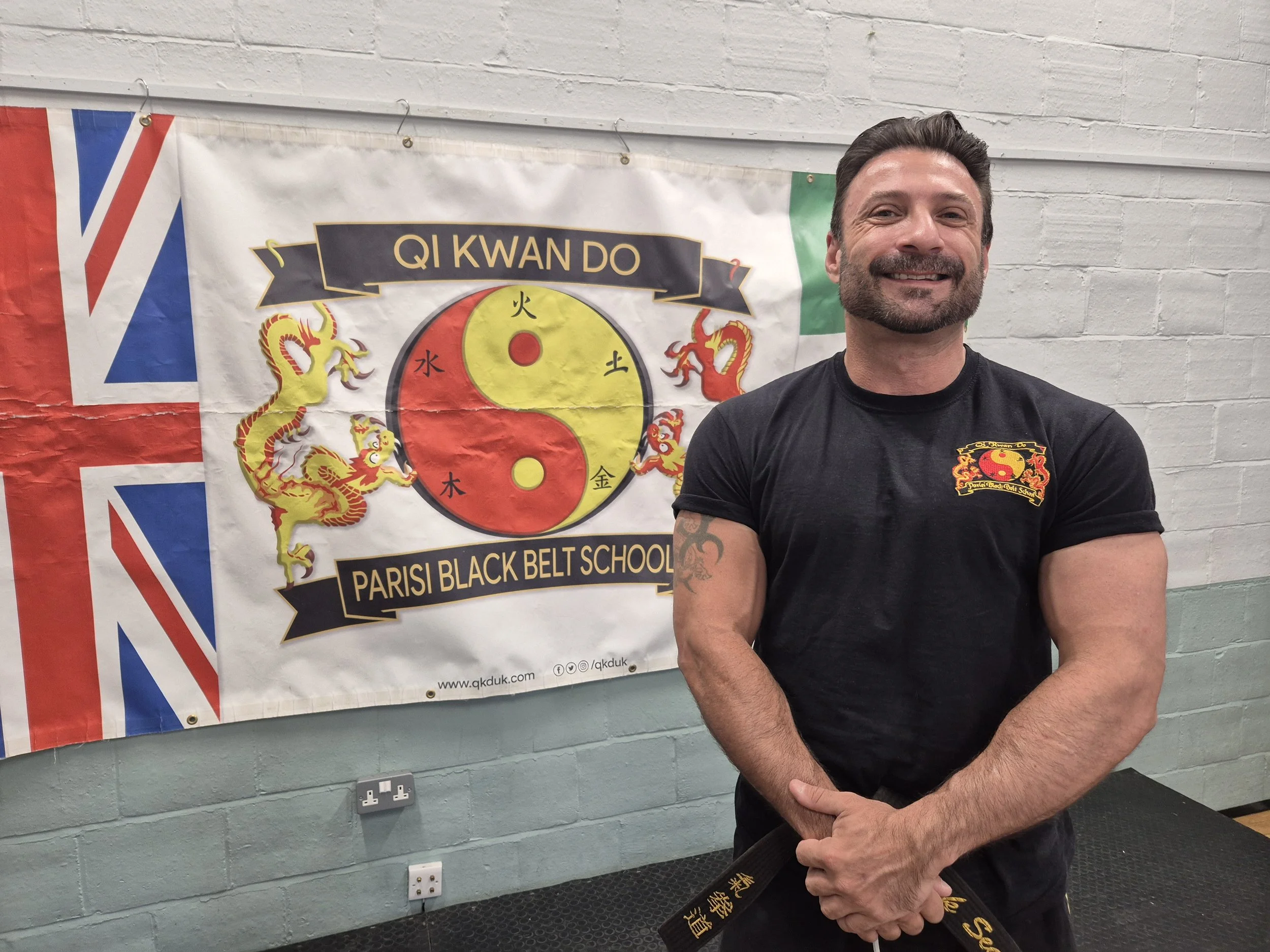 A man with a beard and black T-shirt with a martial arts logo on the sleeve is smiling and standing in front of a banner that reads 'QI KWAN DO' and 'Parisi Black Belt School' with Chinese symbols, a yin-yang symbol, and dragon illustrations. The ban