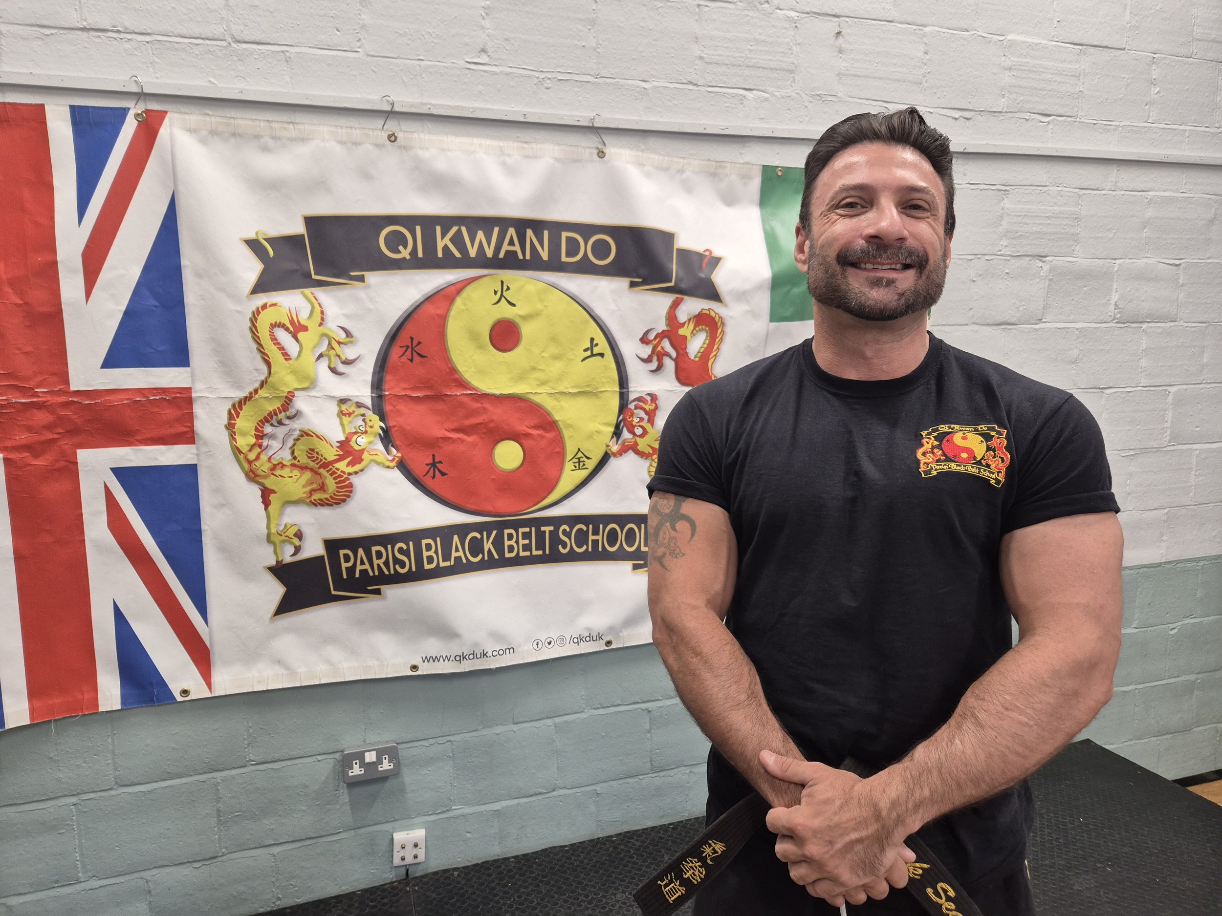 A man with dark, styled hair and a beard smiling, wearing a black t-shirt with a martial arts emblem, standing in front of a martial arts school banner with Chinese characters, dragons, and a yin-yang symbol, against a background of a white brick wal
