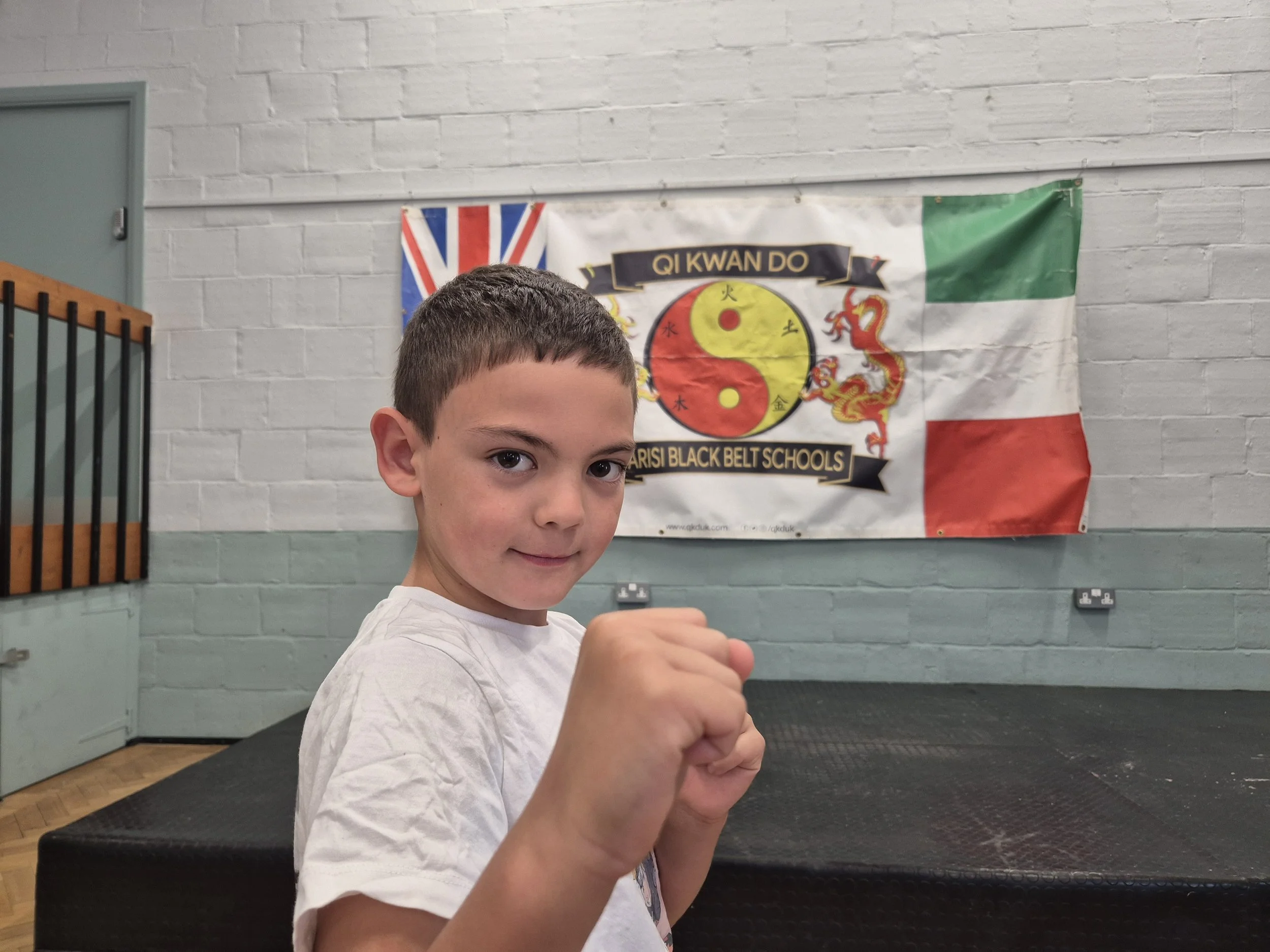Young boy practicing martial arts in a dojo, standing in a fighting stance with fists raised, with a martial arts banner in the background.