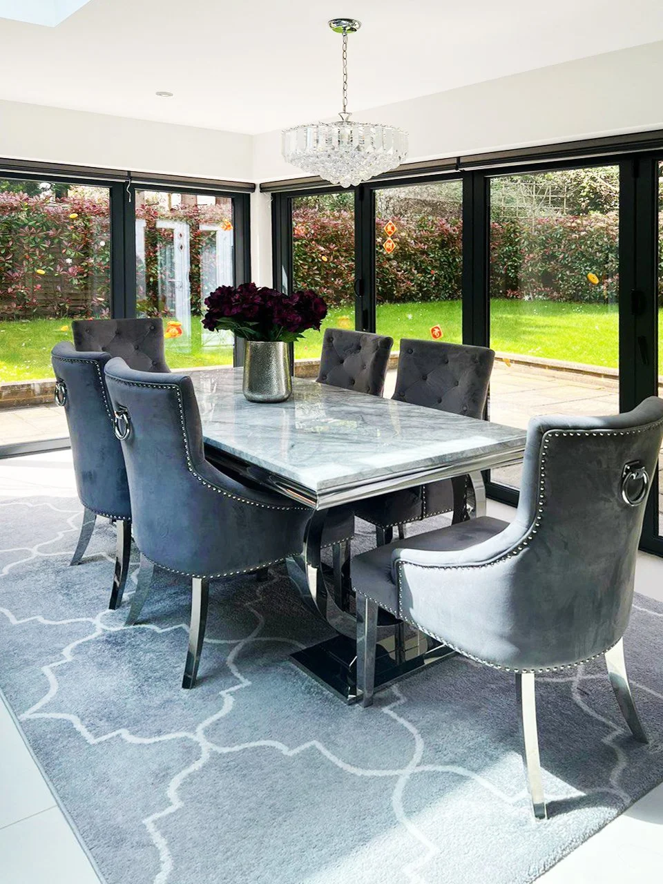 Ein helles Esszimmer mit einem weißen Marmortisch, sechs grauen Samtstühlen, einer Vase mit dunkelroten Blumen, einer Kristalllampe und großen Glasfenstern mit Blick auf einen Garten mit Büschen und Gras.