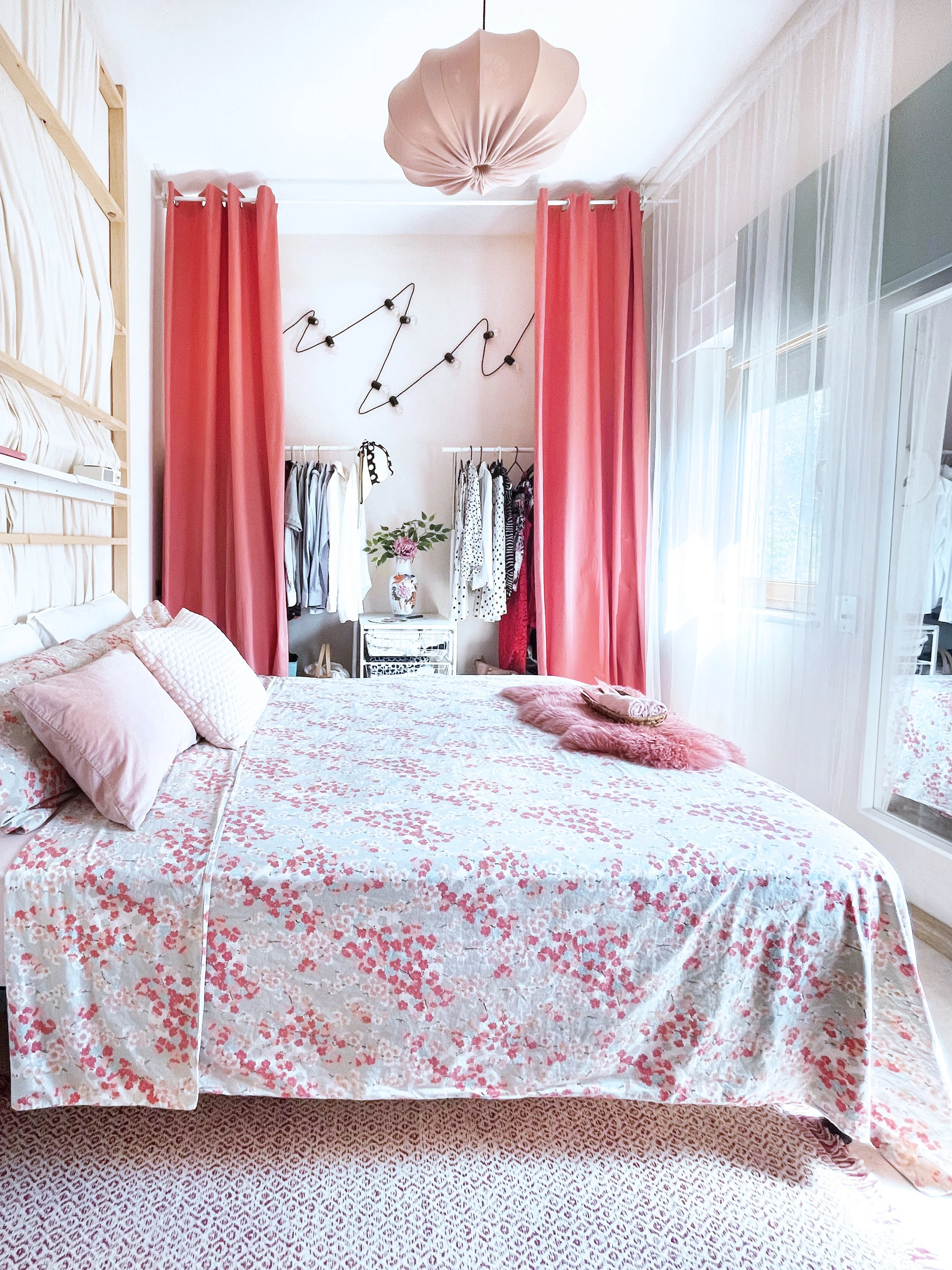 Bett mit floral gemustertem Bettbezug in Weiß und Rosa, mehrere Kissen und eine Decke mit Fell-Optik. Im Hintergrund Kleiderstange mit Kleidung, vordere rote Vorhänge, Hängelampe mit rosa Stoffüberzug, weiße Wand mit Wanddeko und eine Vase mit Blumen