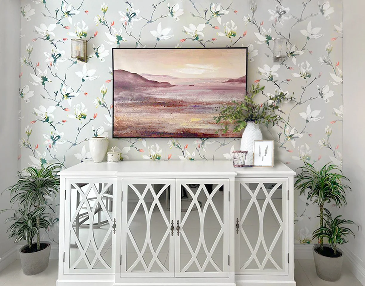 Ein weißes Sideboard vor einer Wand mit Blumenkontrast. Auf dem Sideboard stehen Vasen, Bilder und Dekoration. Darum zwei Pflanzen in Töpfen.