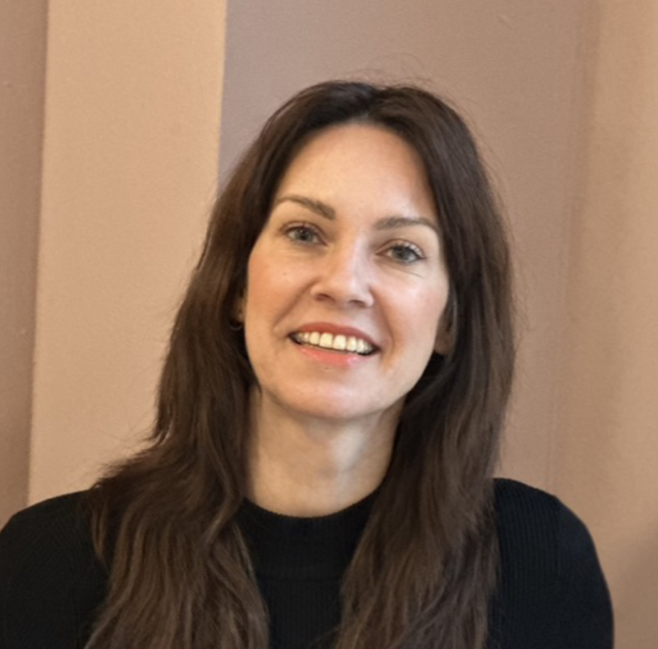 A woman with dark, long hair and a friendly smile, wearing a black sweater, against a beige background.