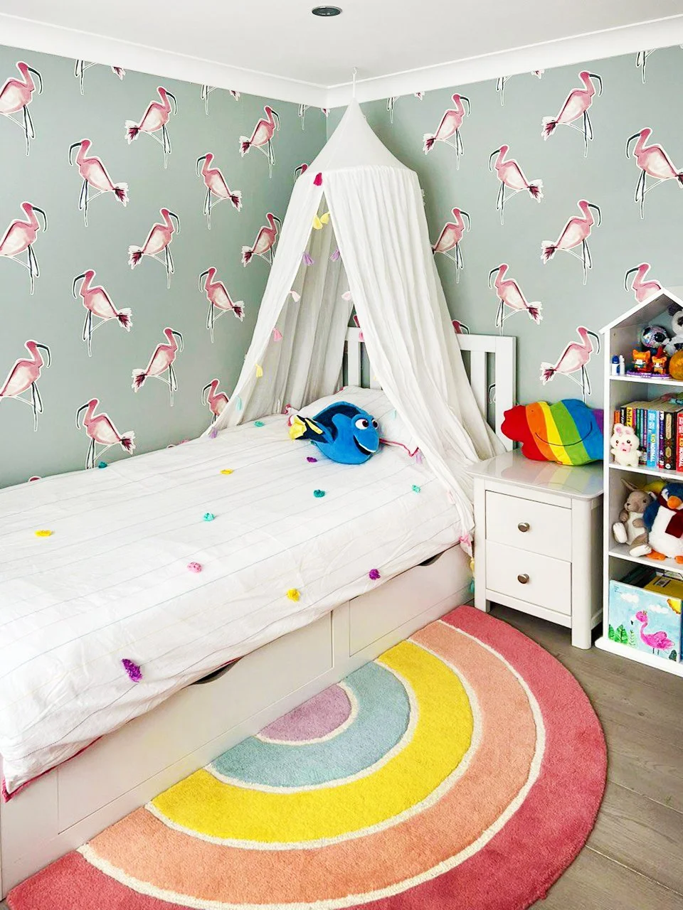 Ein Kinderzimmer mit Flamingo-Tapete, einem weißen Bett mit Vorhang und bunten Dekorationen, einem Sammelregal mit Spielzeug und Büchern, und einem bunten Regenbogen-Teppich auf Holzfußboden.