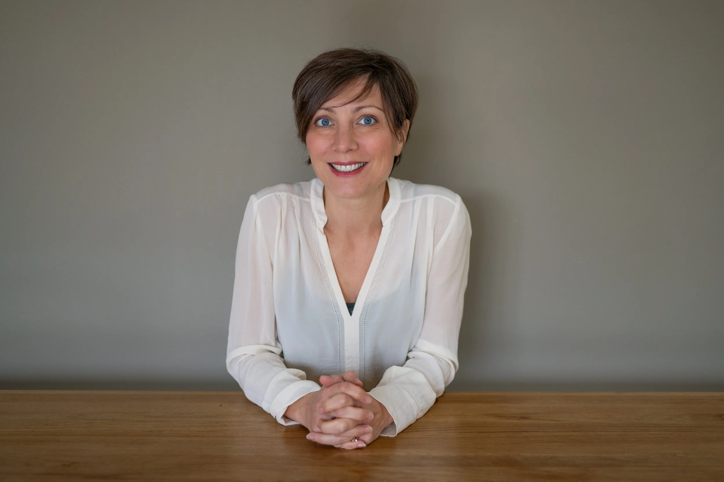 A woman sits at a table in front of a grey wall, wearing a white shirt and smiling at the camera.