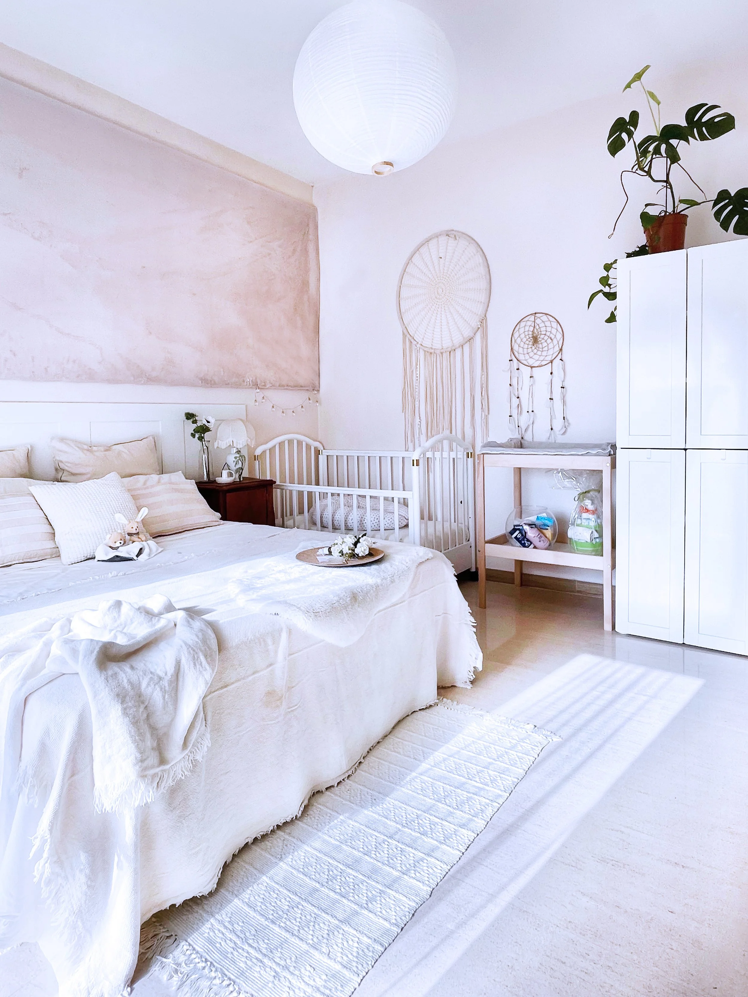 Heller, gemütlicher Schlafzimmerraum mit weißem Bett, Landschaftsdeko, Wickelkommode, hohen weißen Schrank, Wanddeko, Pflanze, weichem Teppich und Papierlaternen Beleuchtung, im Boho-Stil gestaltet.