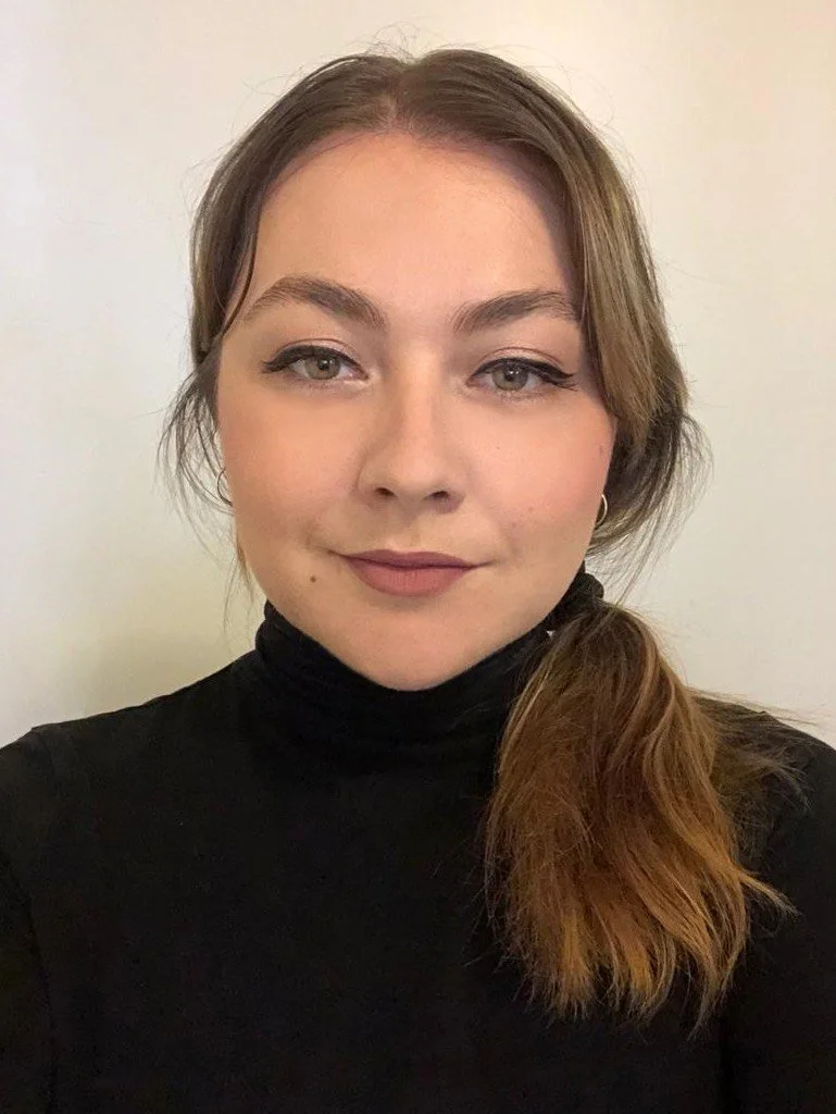 Close-up of a young girl with brown, wavy hair wearing a black turtleneck sweater against a white background