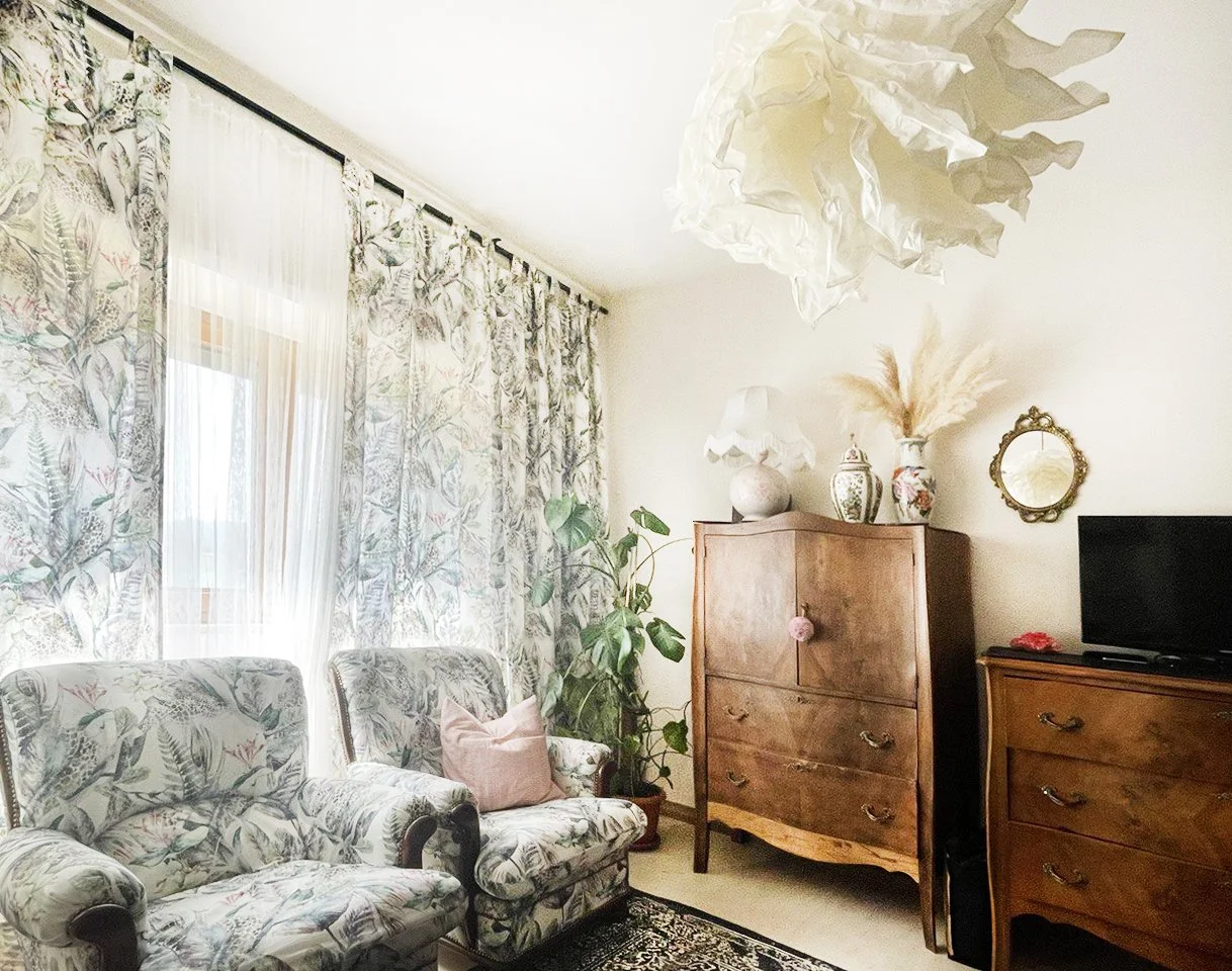 Ein Wohnzimmer mit zwei gepolsterten Sesseln mit Blätter- und Tiermustern, einer topiven Kommode mit Vasen und Lampen, einem kleinen Fernseher, einem Wandspiegel, einem Teppich und Vorhängen mit Blättermuster.