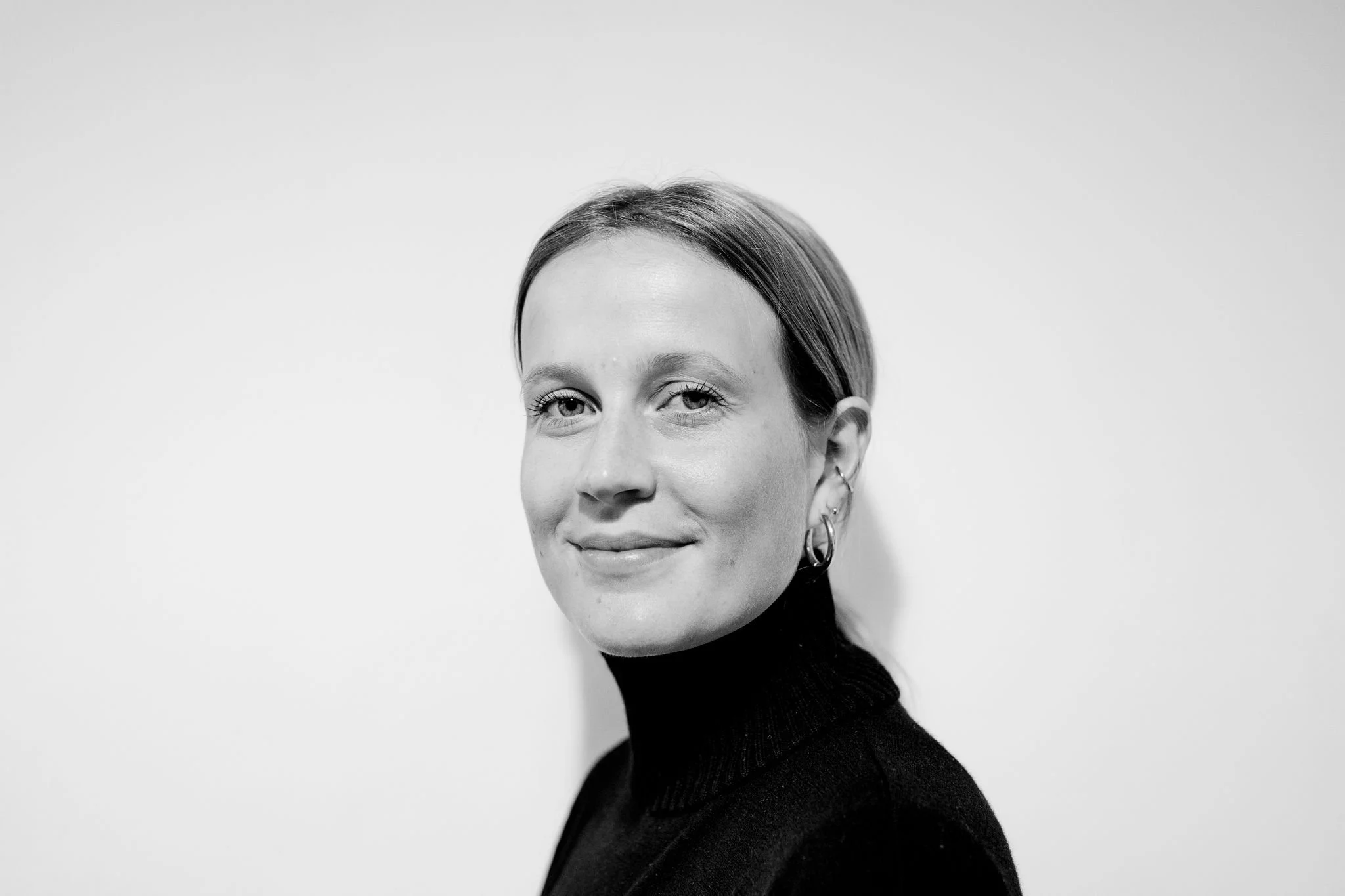 Black and white portrait of a young woman with short hair, several ear piercings and a black turtleneck, against a light background