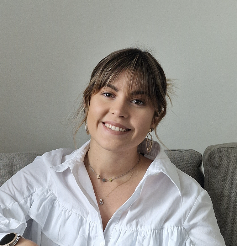 A smiling young girl with brown hair and a white shirt is sitting on a grey sofa.