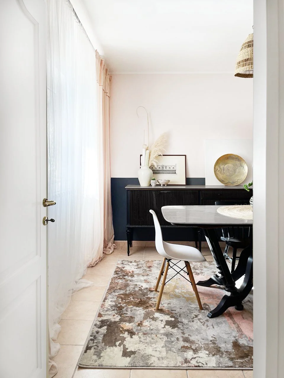 Ein helles Esszimmer mit einem weißen Stuhl, einem schwarzen Tisch und einem schwarzen Sideboard. Dekorative Gegenstände wie eine Vase mit Pampasgras und einen Goldteller stehen auf dem Sideboard. Es gibt weiße, flauschige Vorhänge vor dem Fenster, e