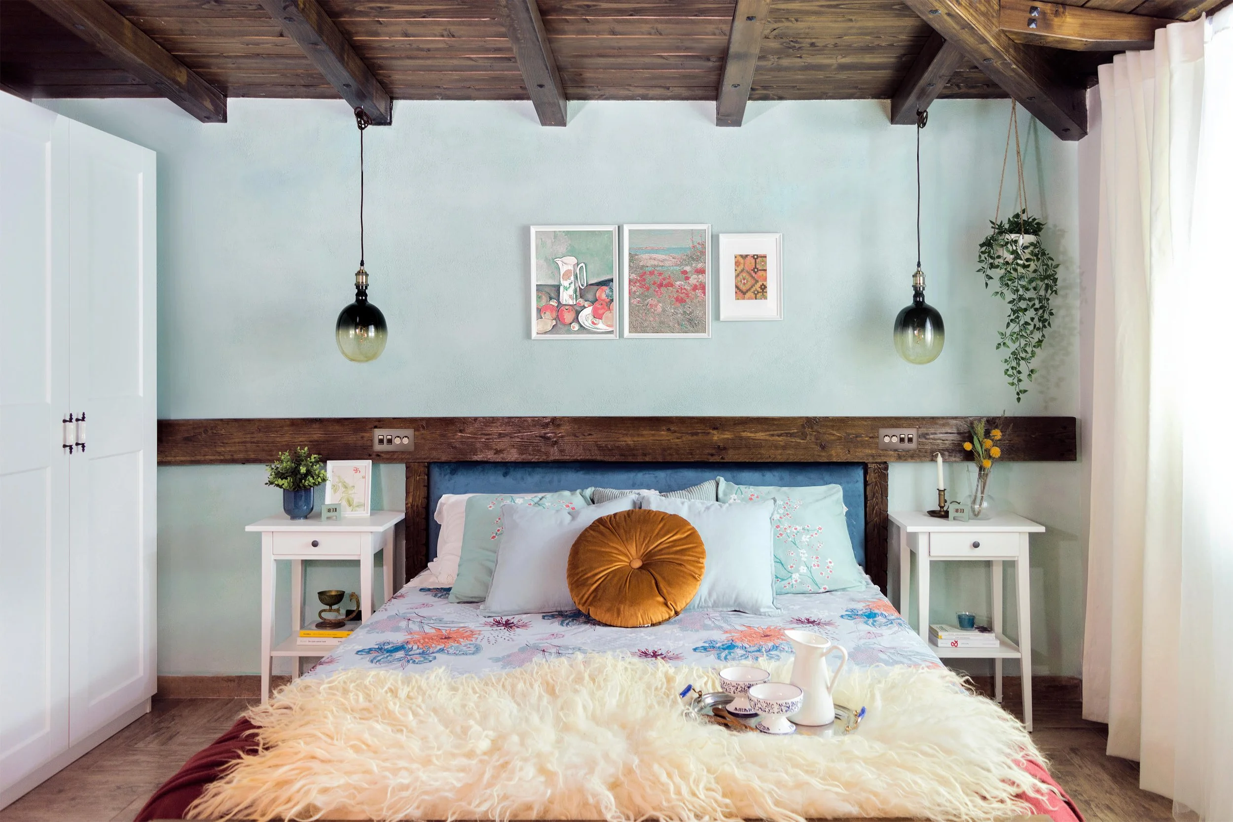 Ein gemütliches Schlafzimmer mit Holzbalken an der Decke, einem Bett mit blumigem Bettzeug, zwei weißen Nachttischen mit Pflanzen und Dekoration, Kunstwerken an der Wand und einer weißen Gardine an einem Fenster.