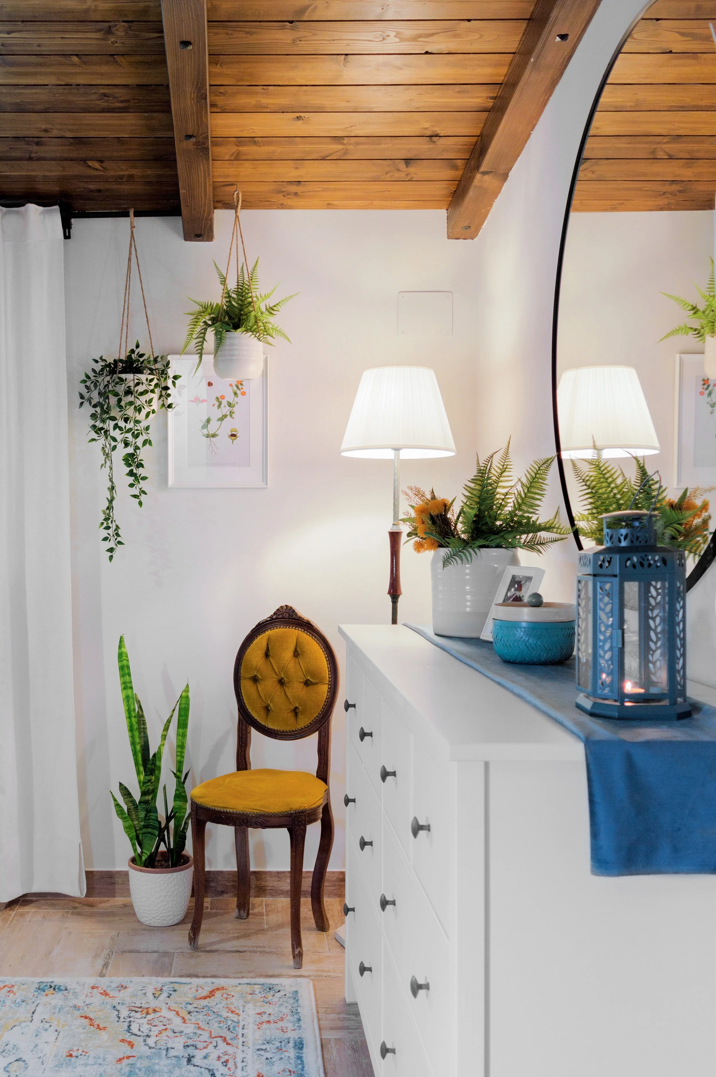 Ein gemütliches Wohnzimmer mit Holzdecke, Pflanzen, einem goldgelben Sessel, einer weißen Schrankkommode, einer Lampe, Dekorationen, einem Spiegel und einem Teppich.