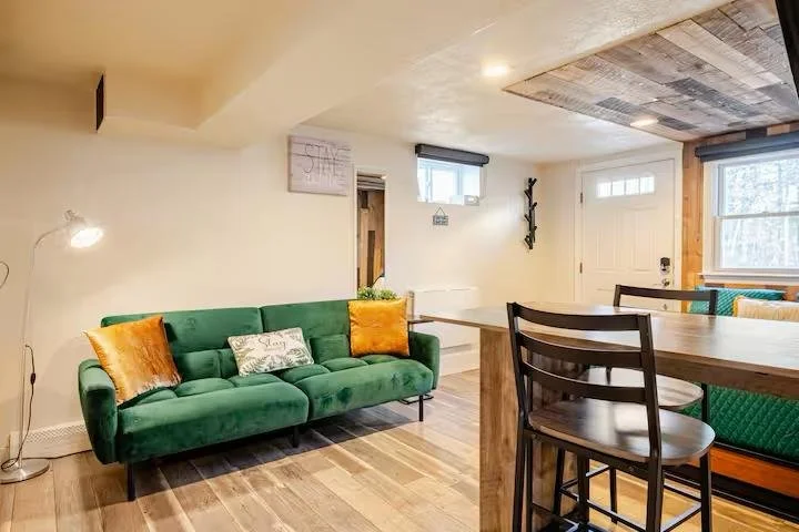Modern basement living area with green velvet sofa, wooden ceiling panel feature, and compact dining setup — interior design with warm tones and natural textures.