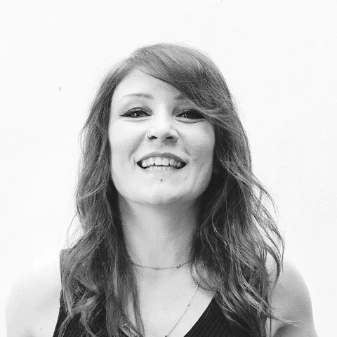 Portrait of a laughing young woman with long, wavy hair and facial piercings, against a white background