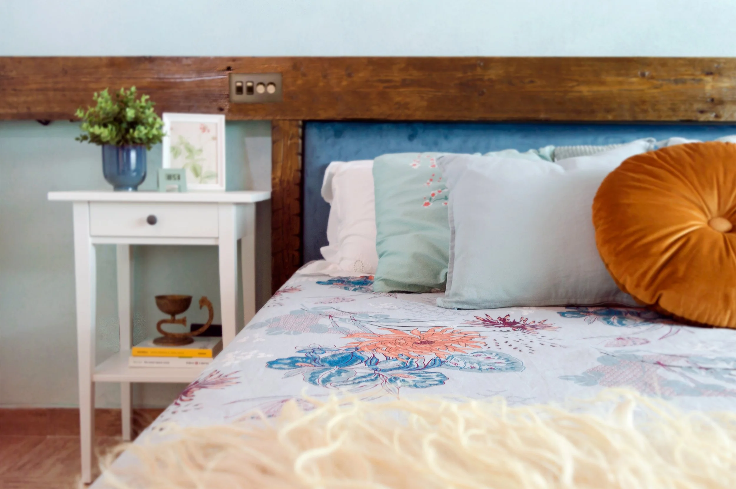 Ein Bett mit bunten Kissen und einer Tagesdecke, daneben ein Nachttisch mit einer Vase mit grünen Blumen, einem Bild und Büchern, an der Wand eine Steckdose.