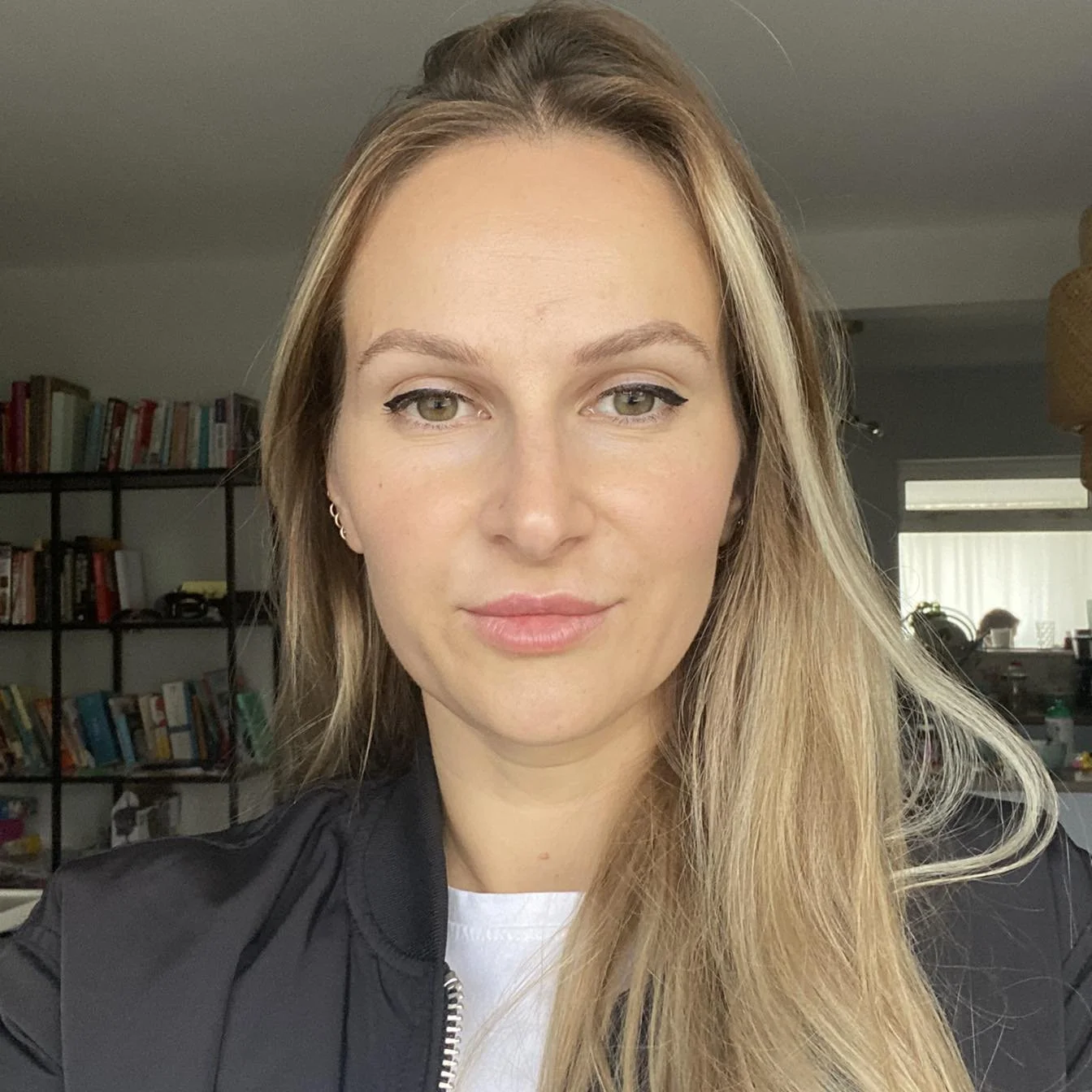 A young girl with long blonde hair, green eyes and subtle makeup, standing in a room with a bookshelf in the background.