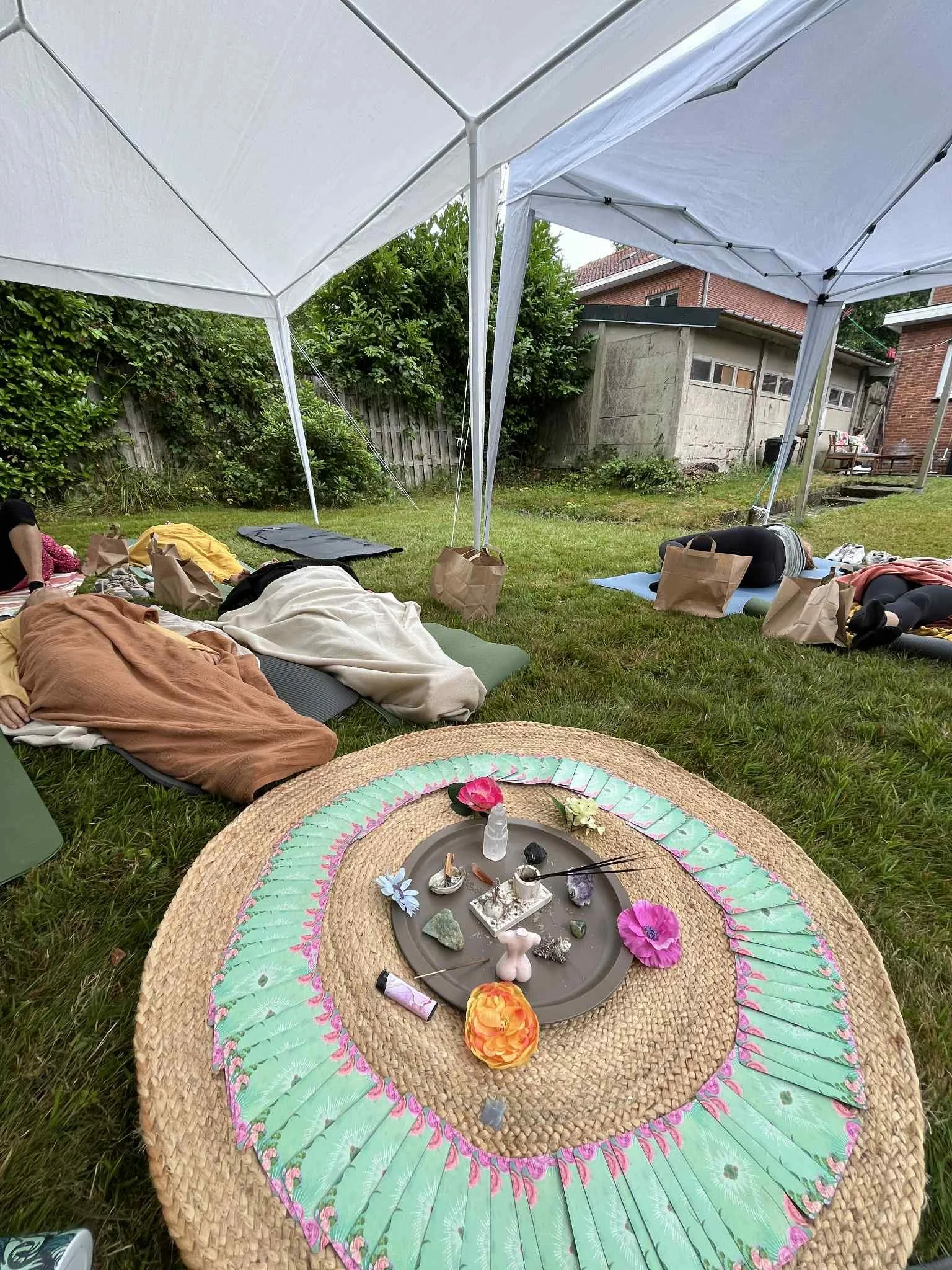Mensen liggen op yoga matten onder een witte tent in een tuin, met een cirkel van tarotkaarten en kristallen op een mat in het midden.