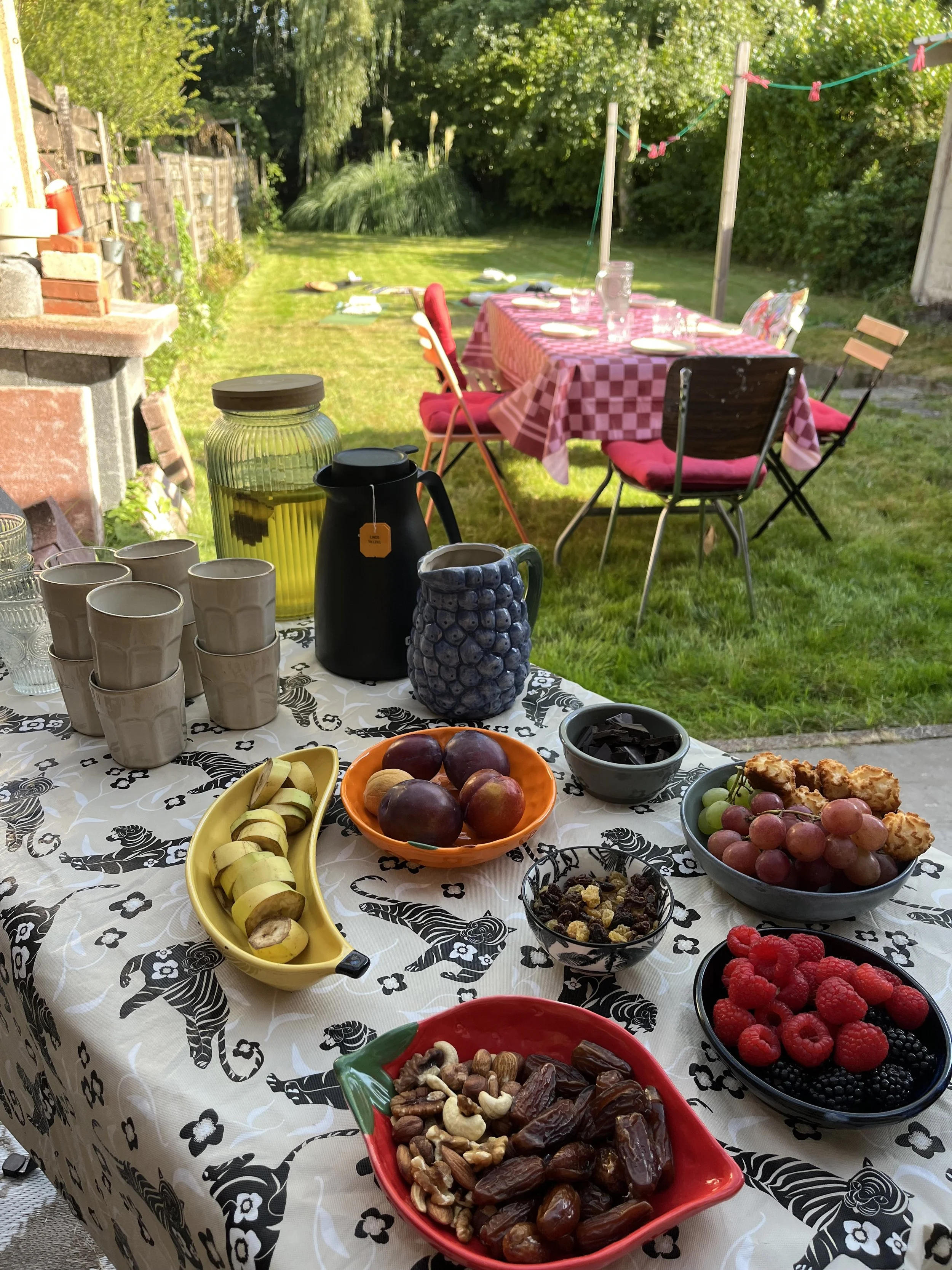 Een tafel met ontbijt zoals diverse kommen met fruit, noten, chocolade, en dranken, buiten in een tuin met een tuinset en ligmatten in de achtergrond.