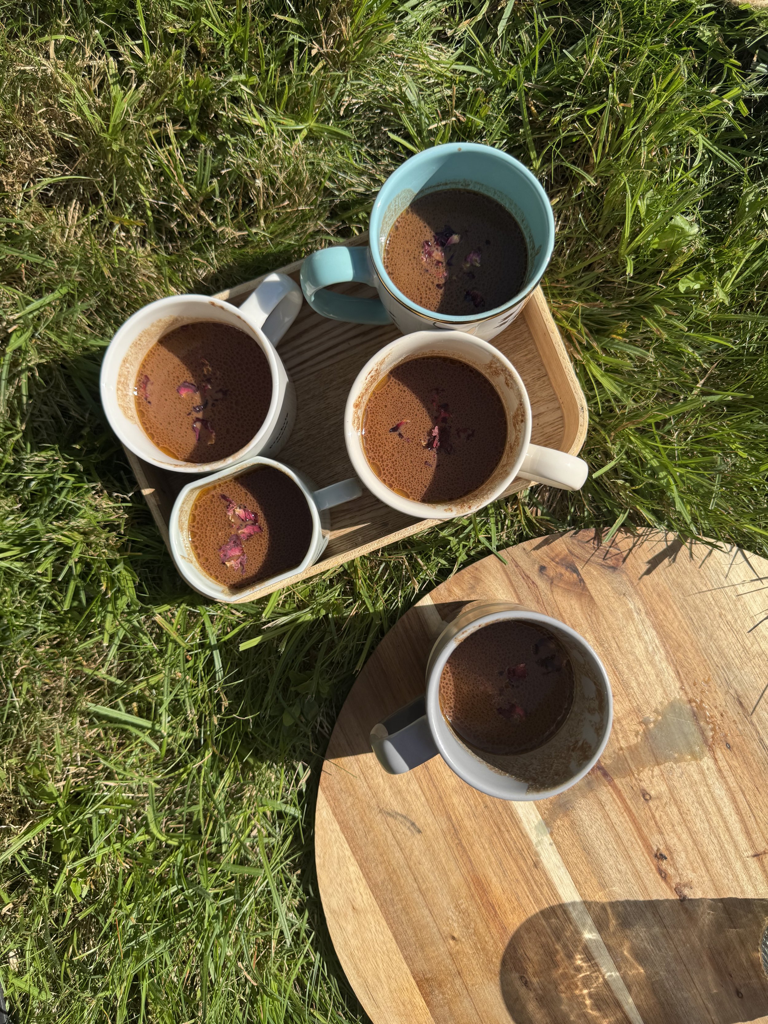 Vijf koppen koffie met bloembladversiering op een houten dienblad en een houten bord, allemaal geplaatst op gras in de zon.