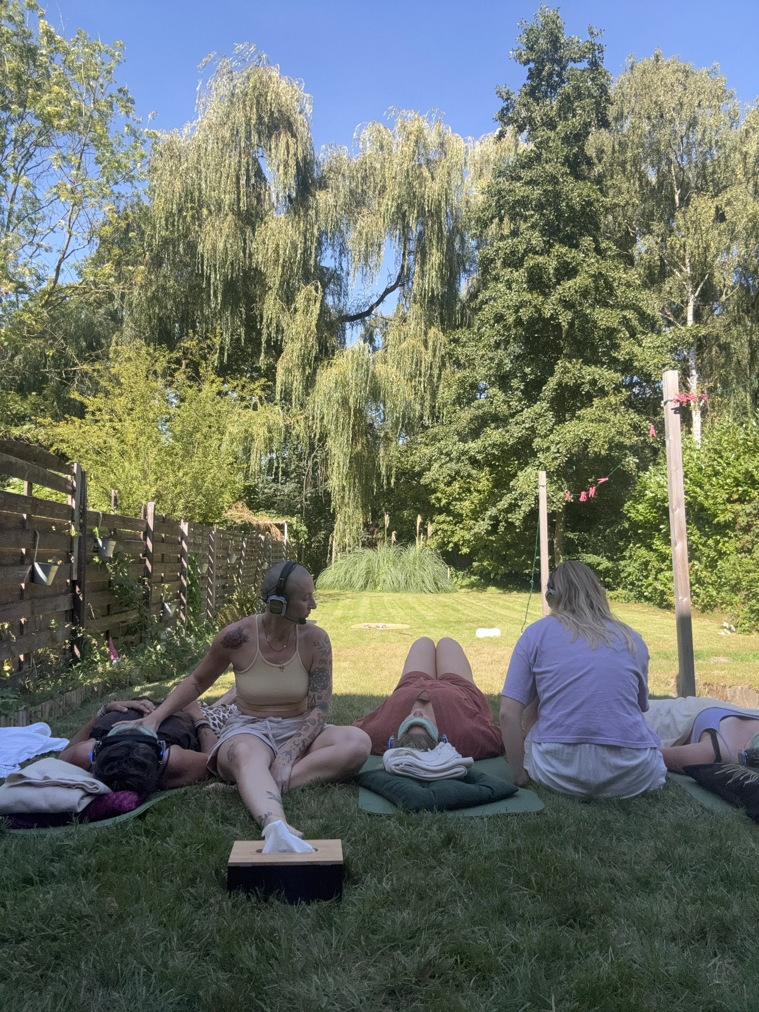 Mensen liggen in een tuin in een zonovergoten omgeving, luisteren naar headsets, met een blauwe lucht, groene bomen en een stuk gras in de achtergrond.
