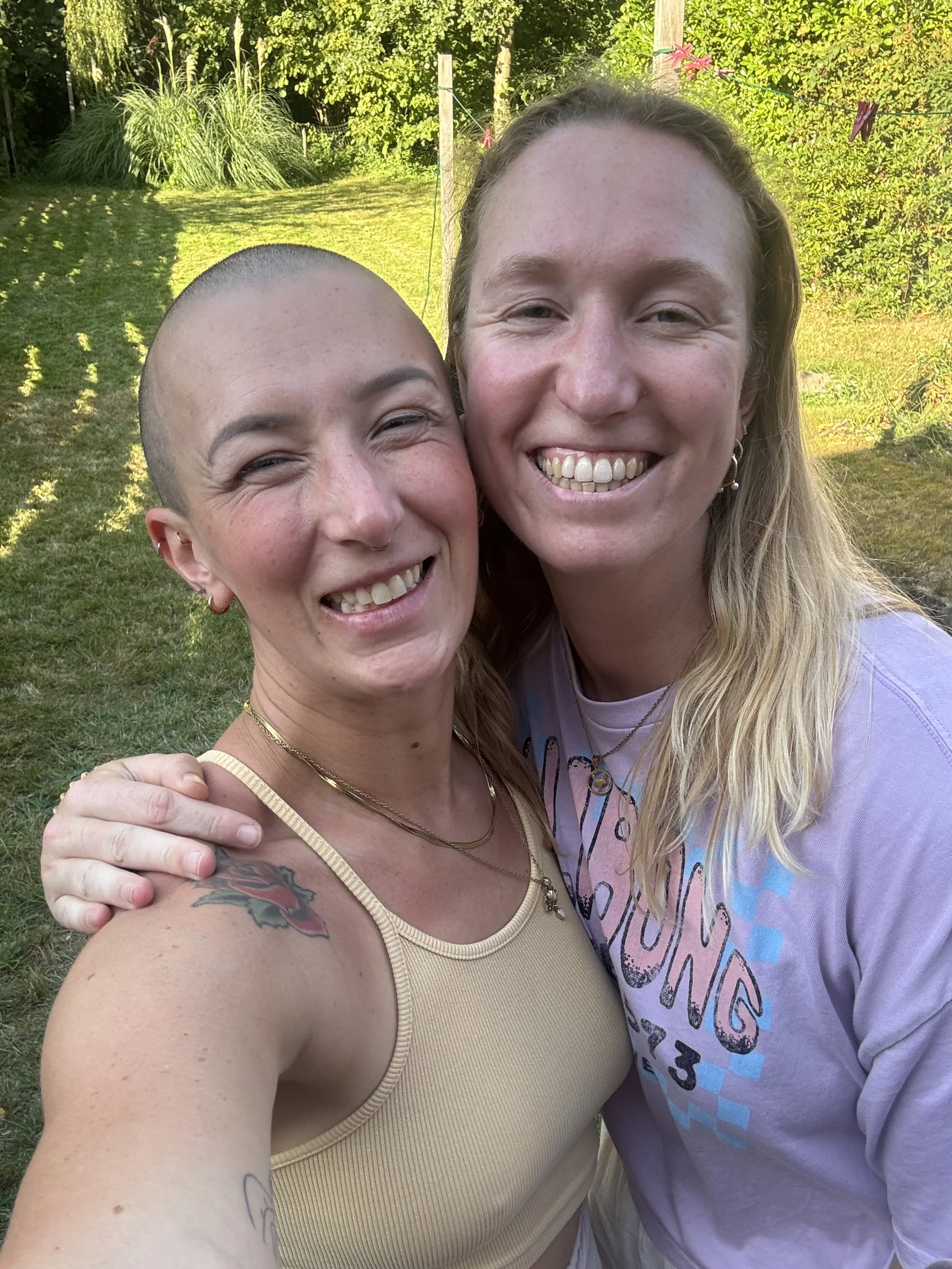 Twee vrouwen die samen glimlachen en poseren voor een tuin, één met een koperen tatoeage op haar schouder, de andere met lang blond haar en een lichte paarse shirt.