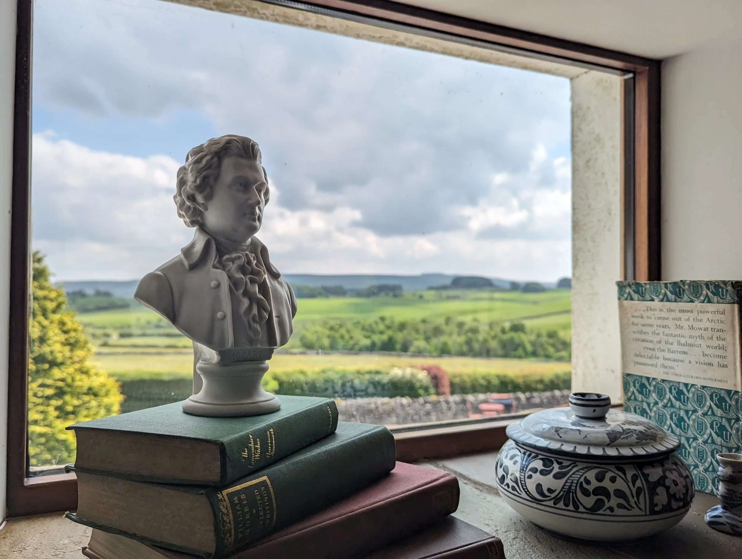 Bust sculpture on stacked books with scenic countryside view through window.