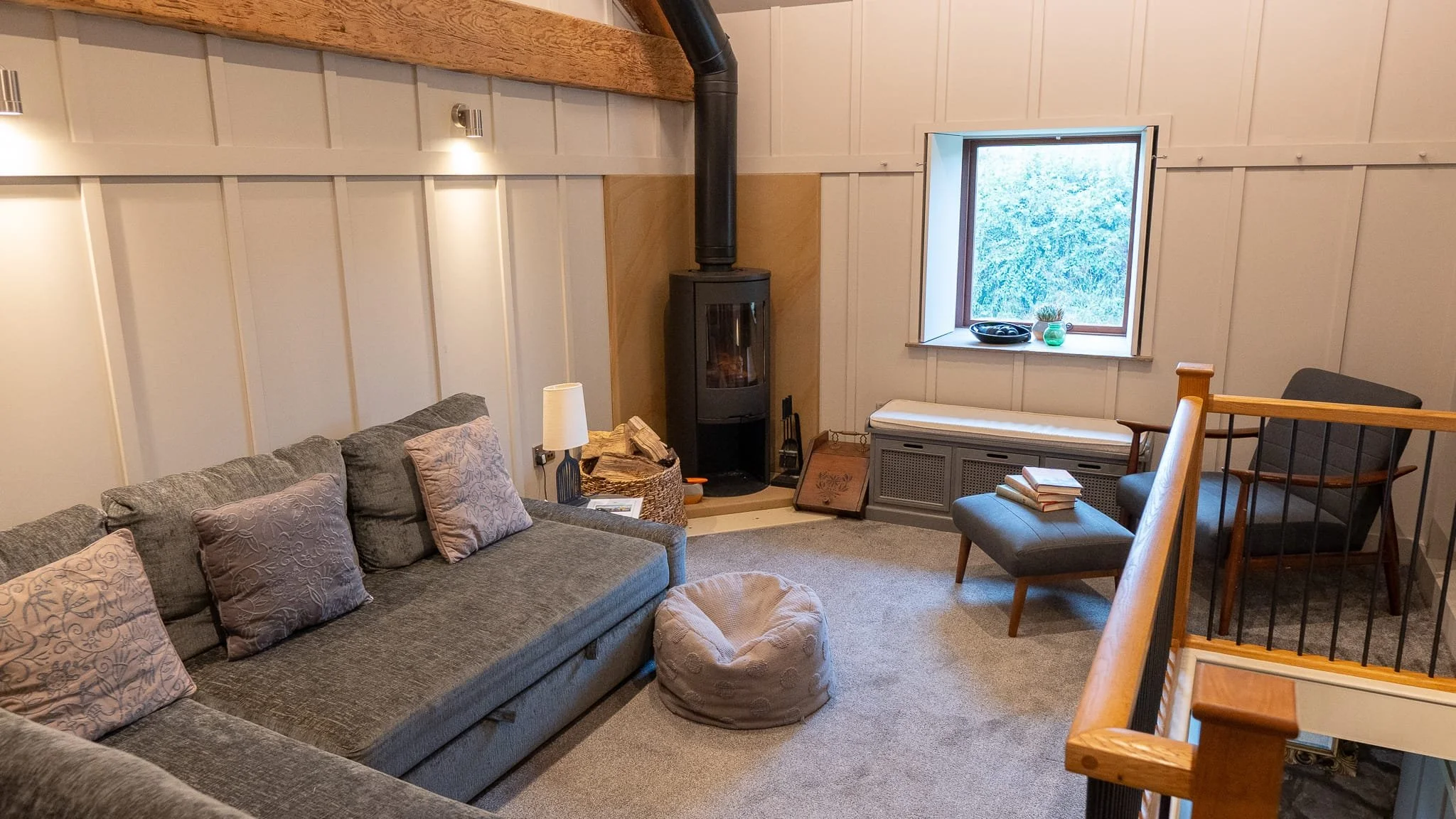 Cozy living room with a gray sectional sofa, decorative pillows, a modern wood-burning stove, and a window seat. There is a cushioned armchair with an ottoman, books, and a bean bag on the carpeted floor. Exposed wooden beams and paneled walls add a rustic touch.